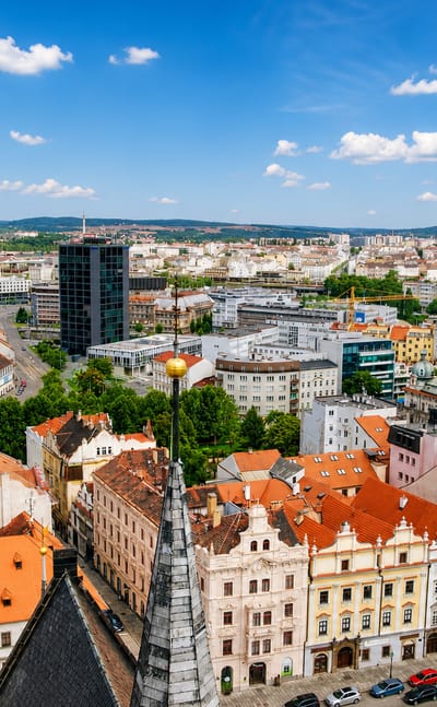 Aerial view of Pilsen's red-roofed old town with St. Bartholomew's spire and a modern tower under blue sky.