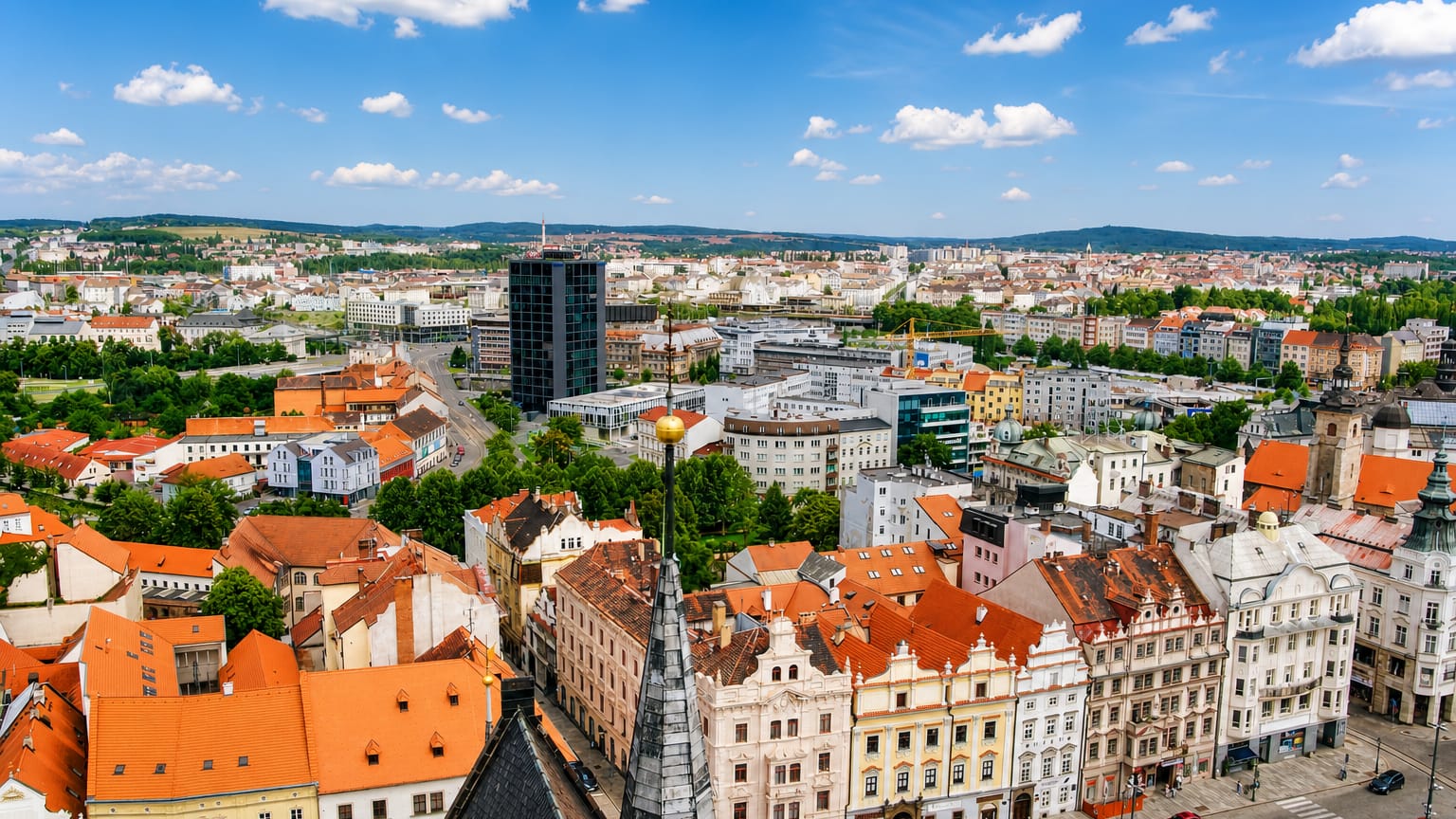 Aerial view of Pilsen's red-roofed old town with St. Bartholomew's spire and a modern tower under blue sky.