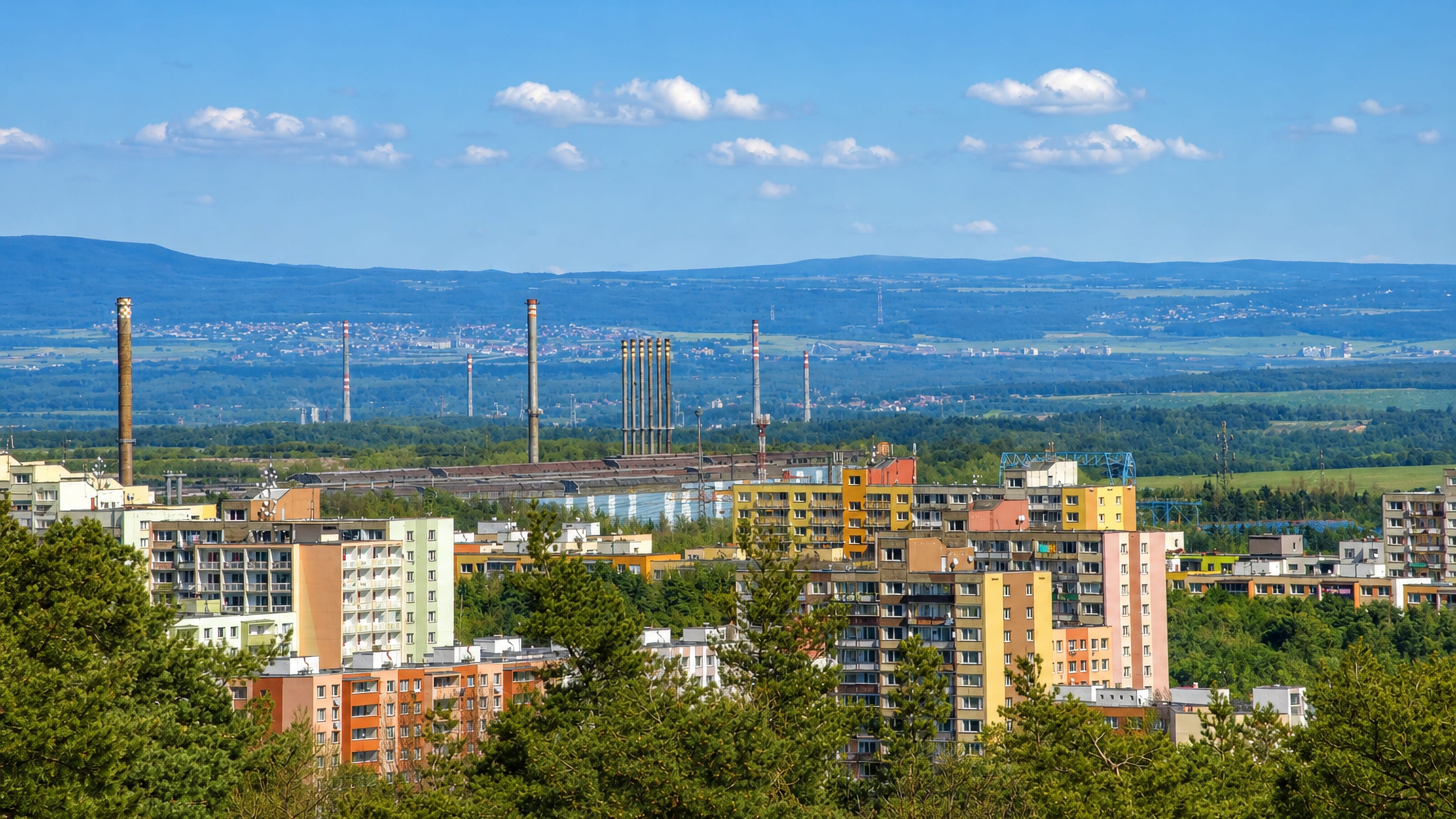 Colourful panel housing blocks among pines in Krocehlavy, Kladno, with industrial chimneys and hills beyond.
