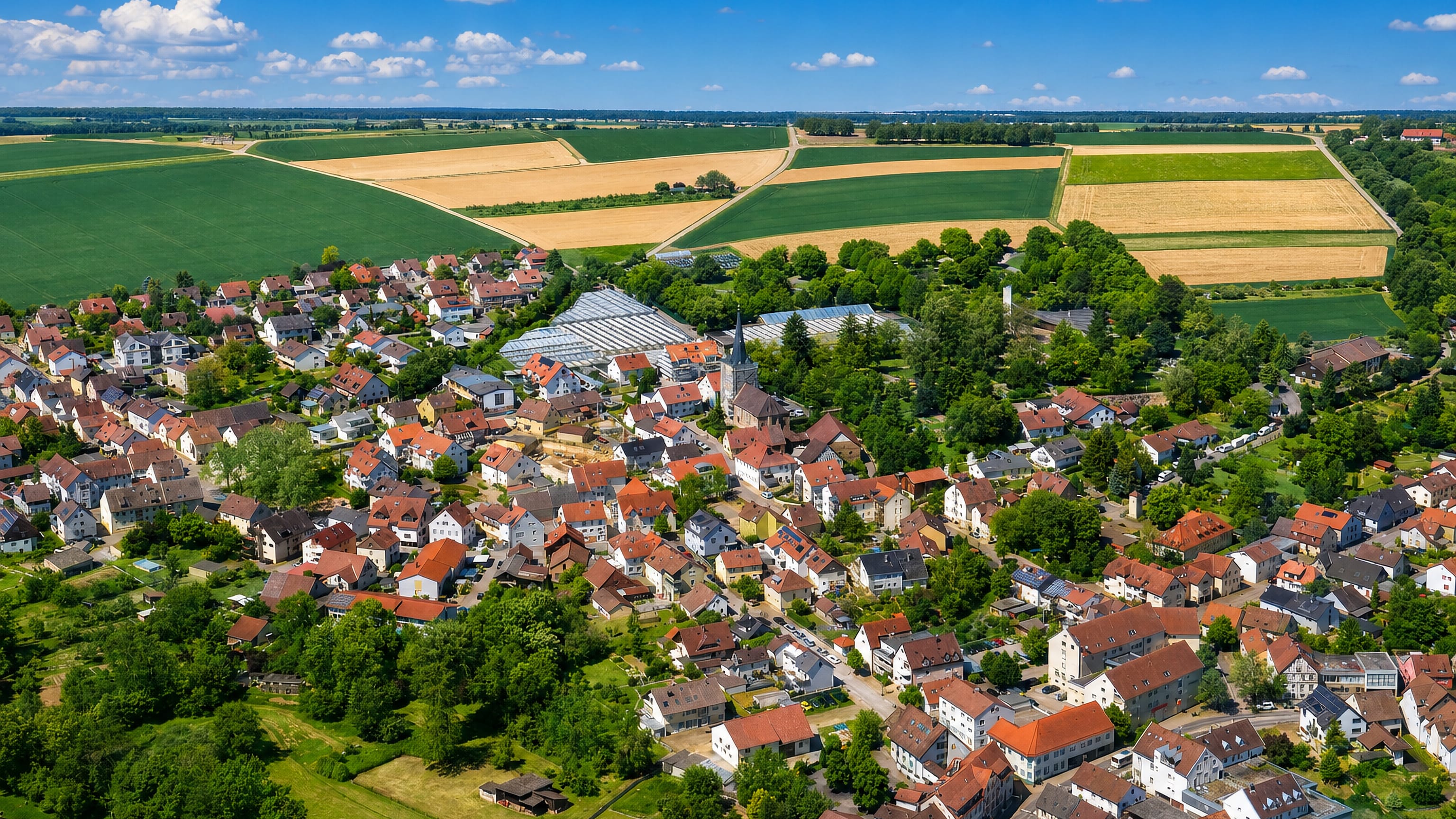 Aerial of Heilbronn-Frankenbach, its church steeple and red roofs ringed by fields and greenhouses.