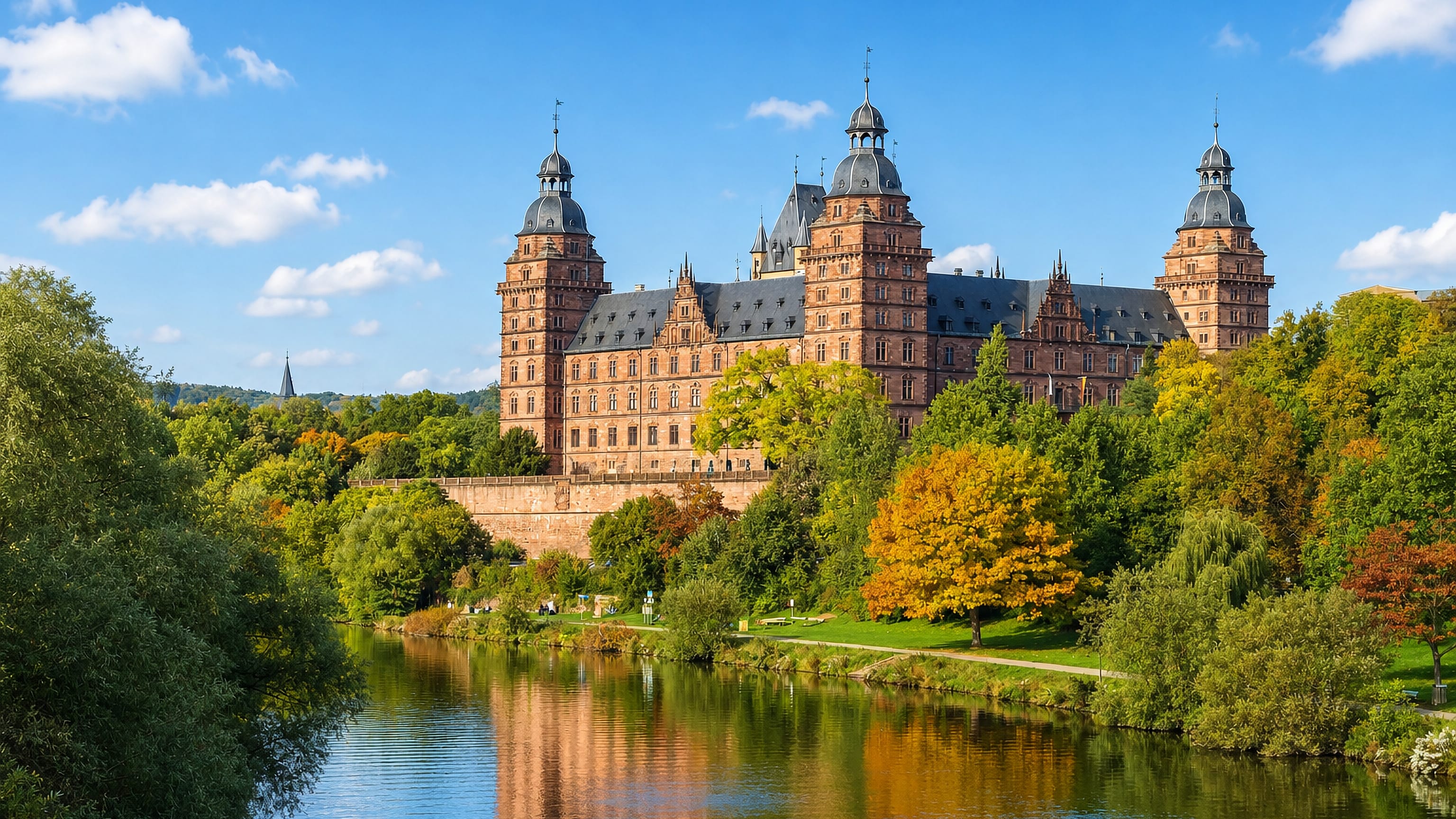 Schloss Johannisburg's red sandstone towers rising above the tree-lined River Main in Aschaffenburg, Bavaria.