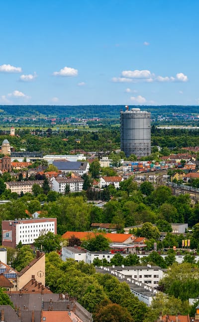 Elevated view across Augsburg's rooftops, churches and a large gasometer with green suburbs beyond.