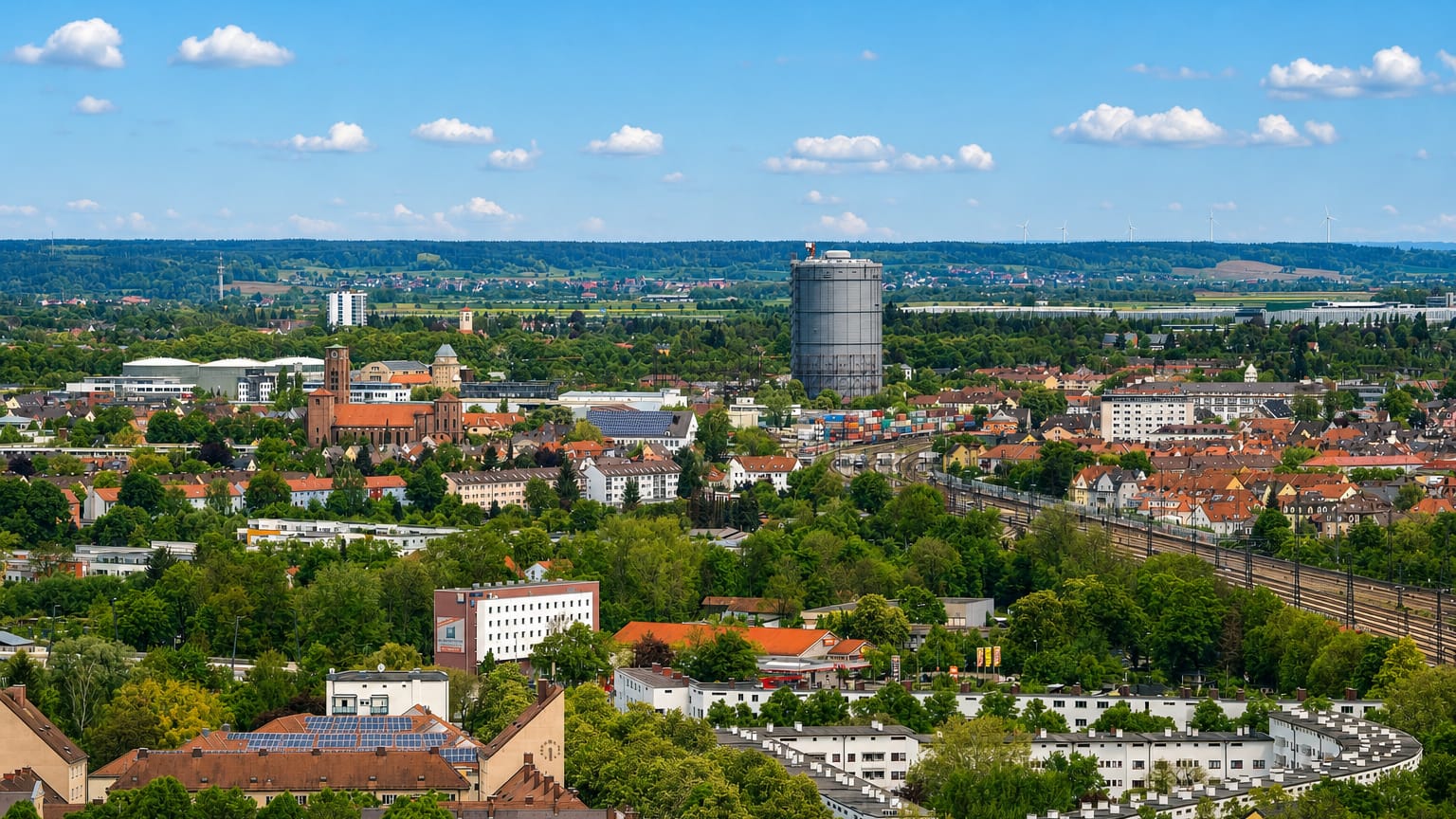 Elevated view across Augsburg's rooftops, churches and a large gasometer with green suburbs beyond.