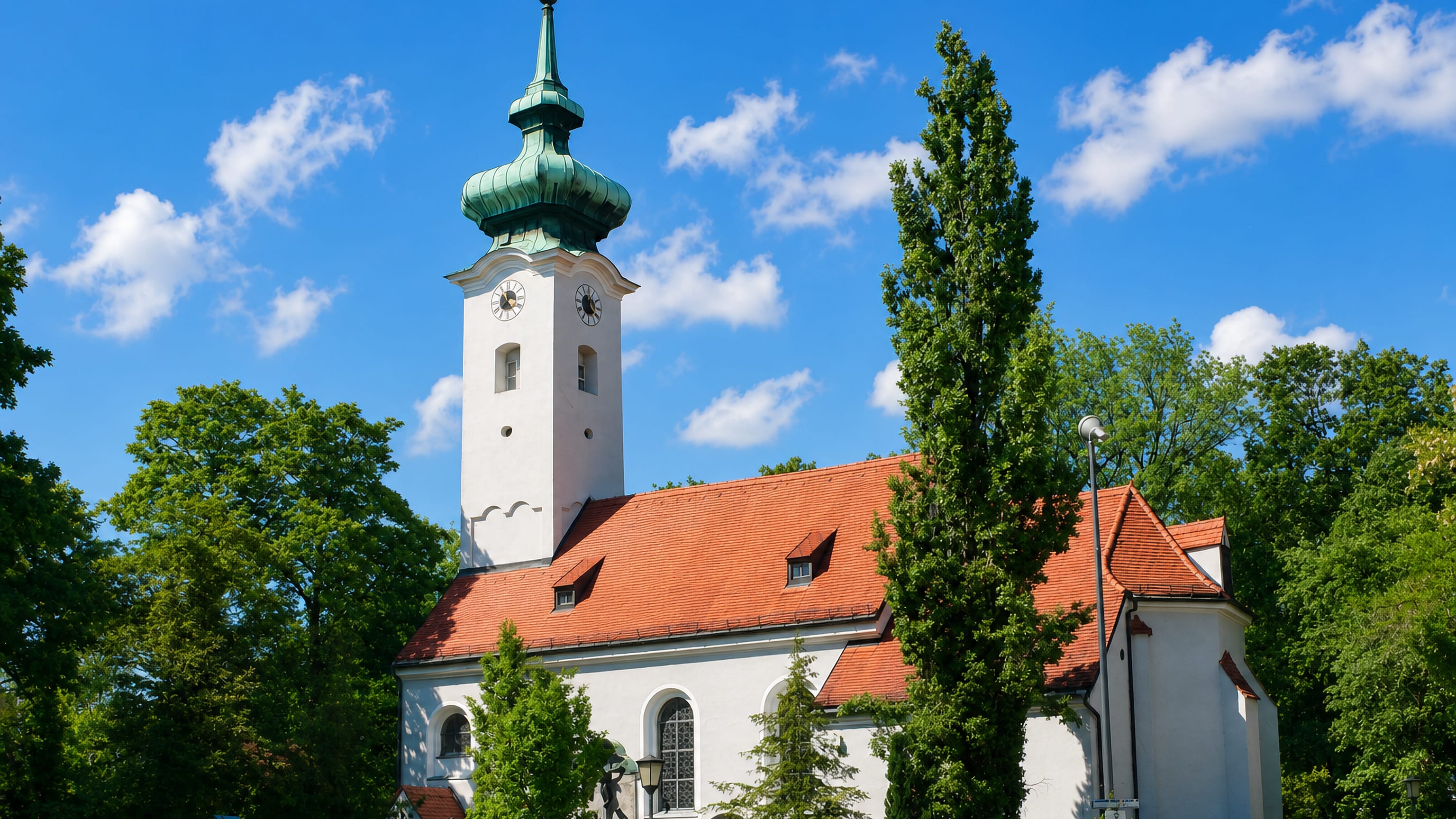 Whitewashed onion-domed parish church with red tile roof among summer trees in Bogenhausen, Munich.