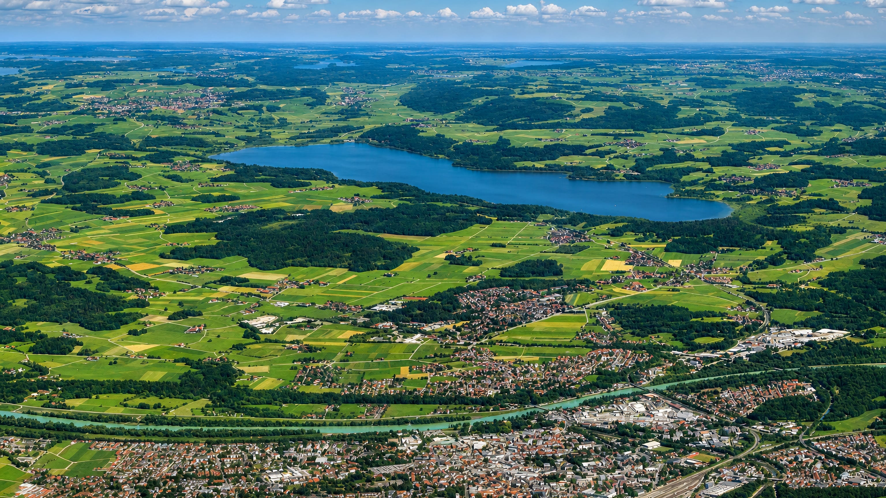 High aerial of Rosenheim beside the turquoise Inn river, with Bavarian farmland and a lake stretching to the horizon.