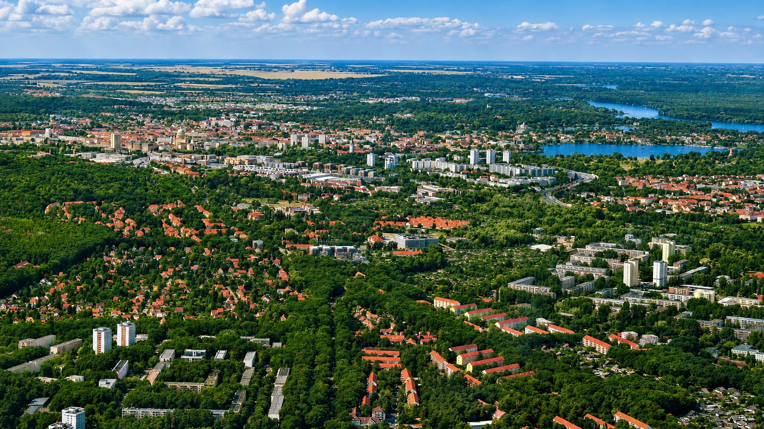 Aerial daytime panorama of Potsdam, red-roofed districts and tower blocks amid greenery beside the Havel lakes.