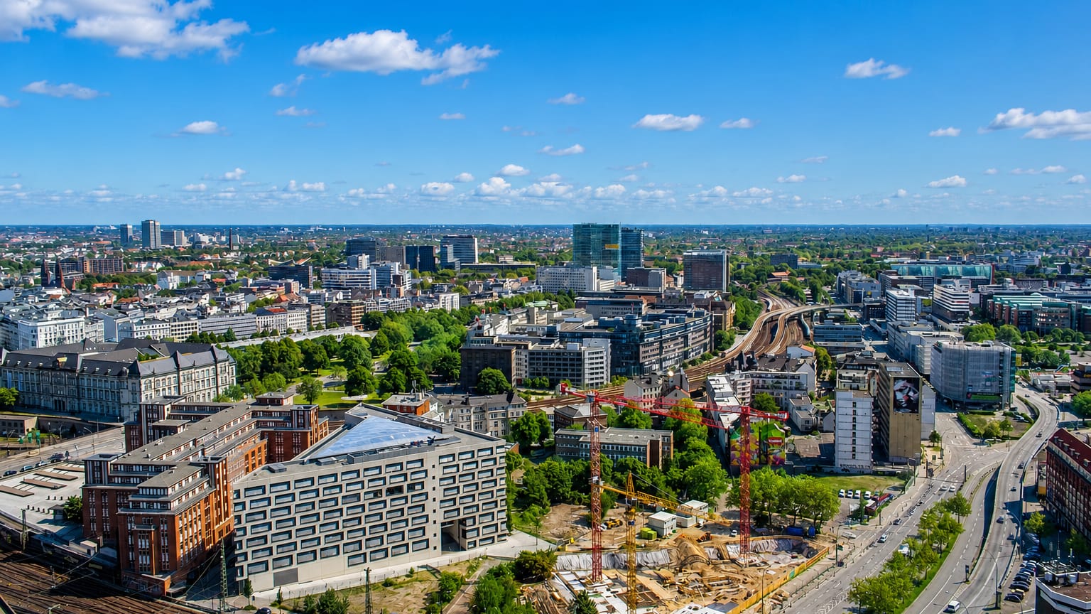 Aerial view over the Hammerbrook district of Hamburg with mid-rise offices, rail lines and construction cranes under blue sky.
