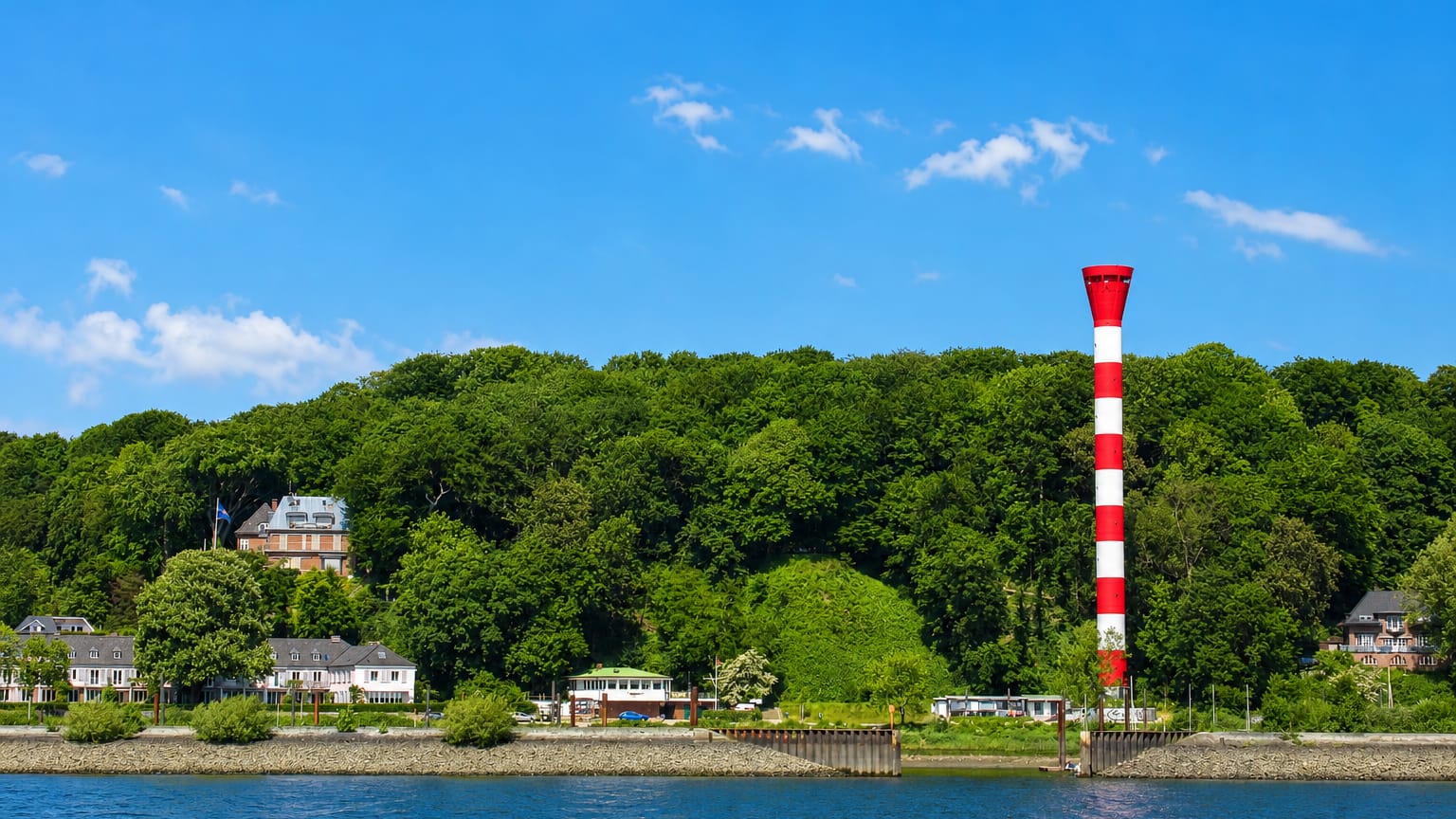The wooded Elbe riverbank at Nienstedten in Hamburg with villas along the shore and a red-and-white striped lighthouse.