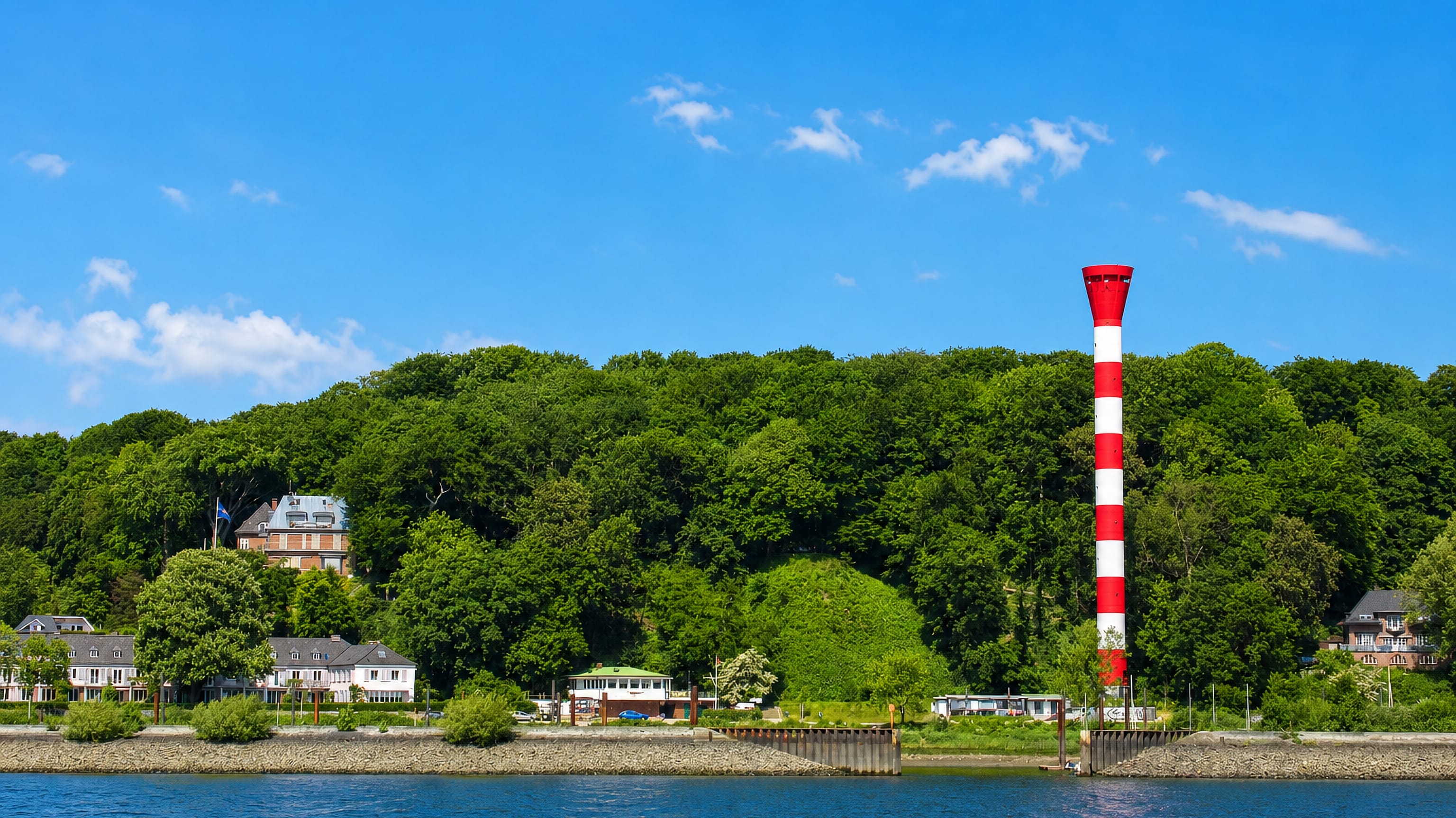 The wooded Elbe riverbank at Nienstedten in Hamburg with villas along the shore and a red-and-white striped lighthouse.