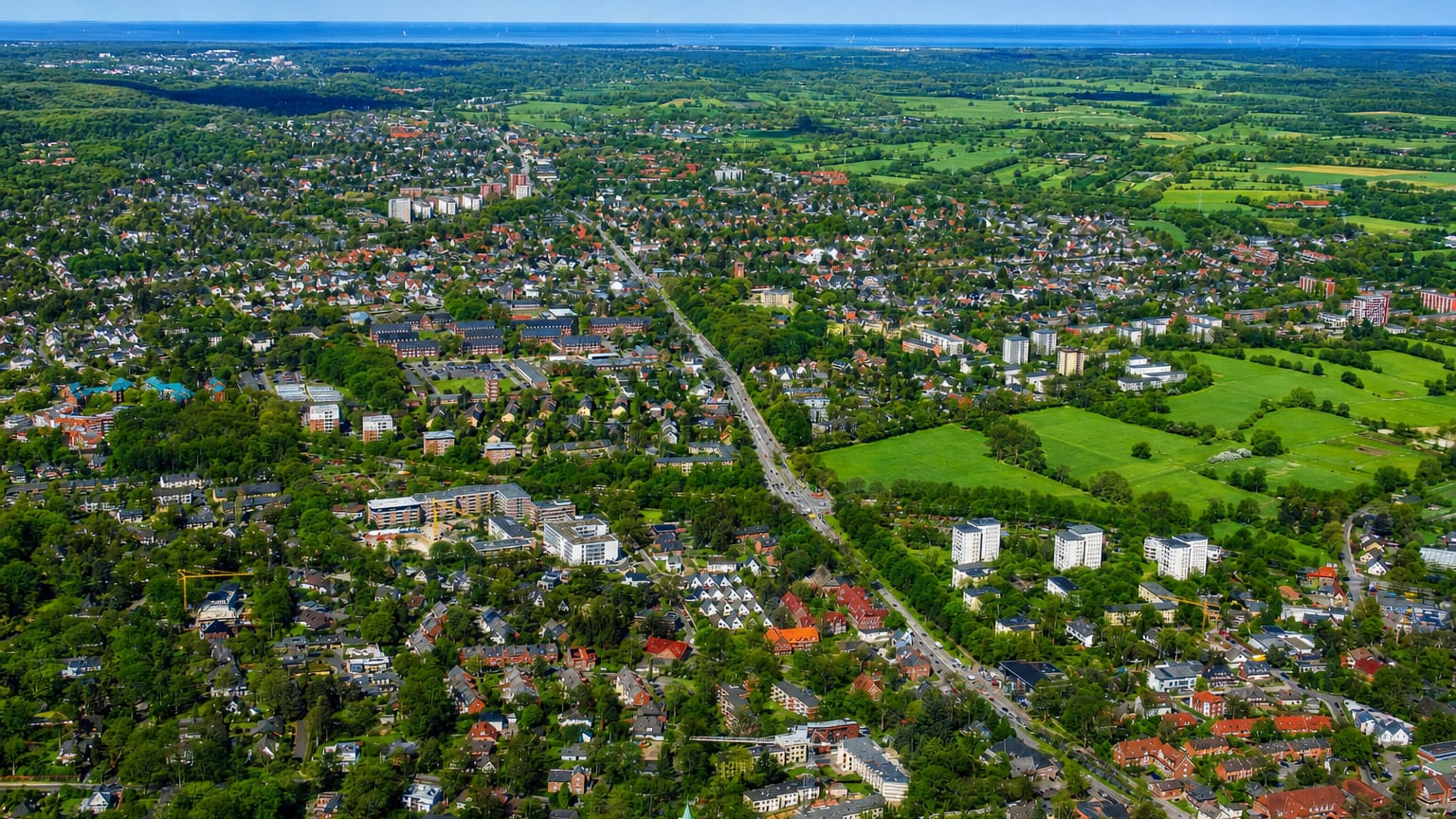 Aerial view of the Osdorf district of Hamburg, low-rise housing and apartment blocks along a road corridor with green fields and distant water.