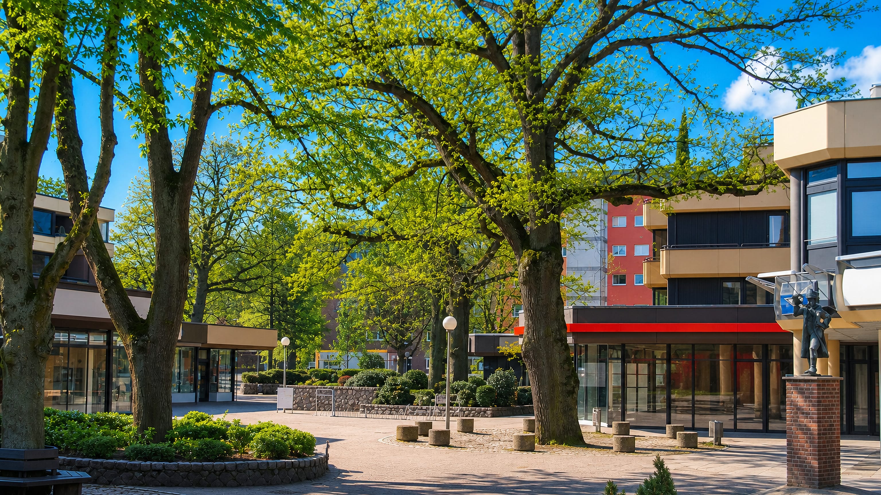 Leafy pedestrian square with low shopfronts, a statue and apartment blocks in Volksdorf, Hamburg.