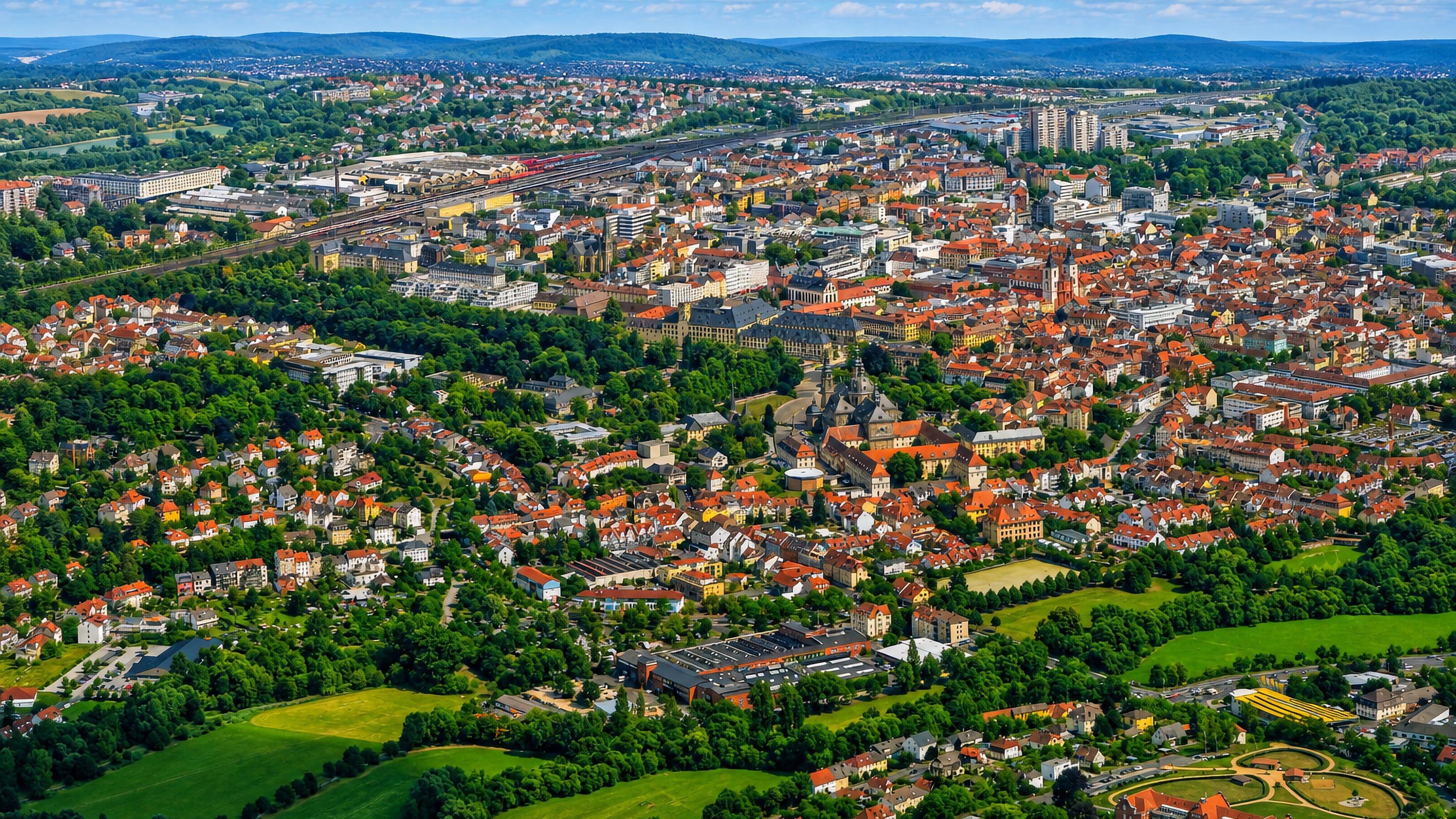 Aerial view over Fulda, Germany, with the baroque cathedral and city palace amid red-roofed streets and green hills.