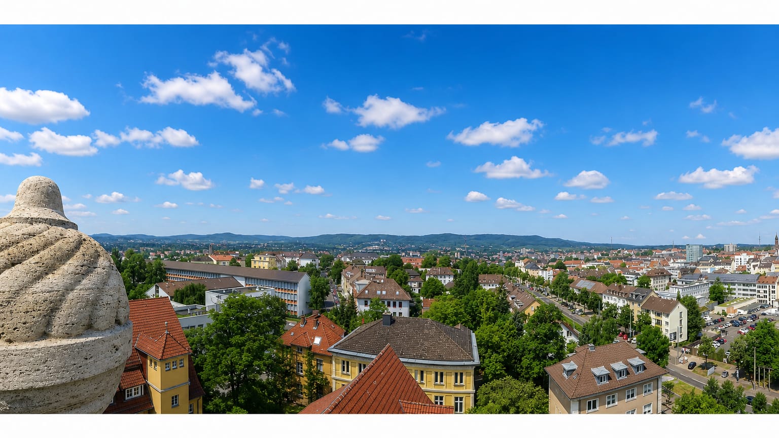 Panorama over Kassel's red rooftops and wooded hills viewed past a stone monument figure.