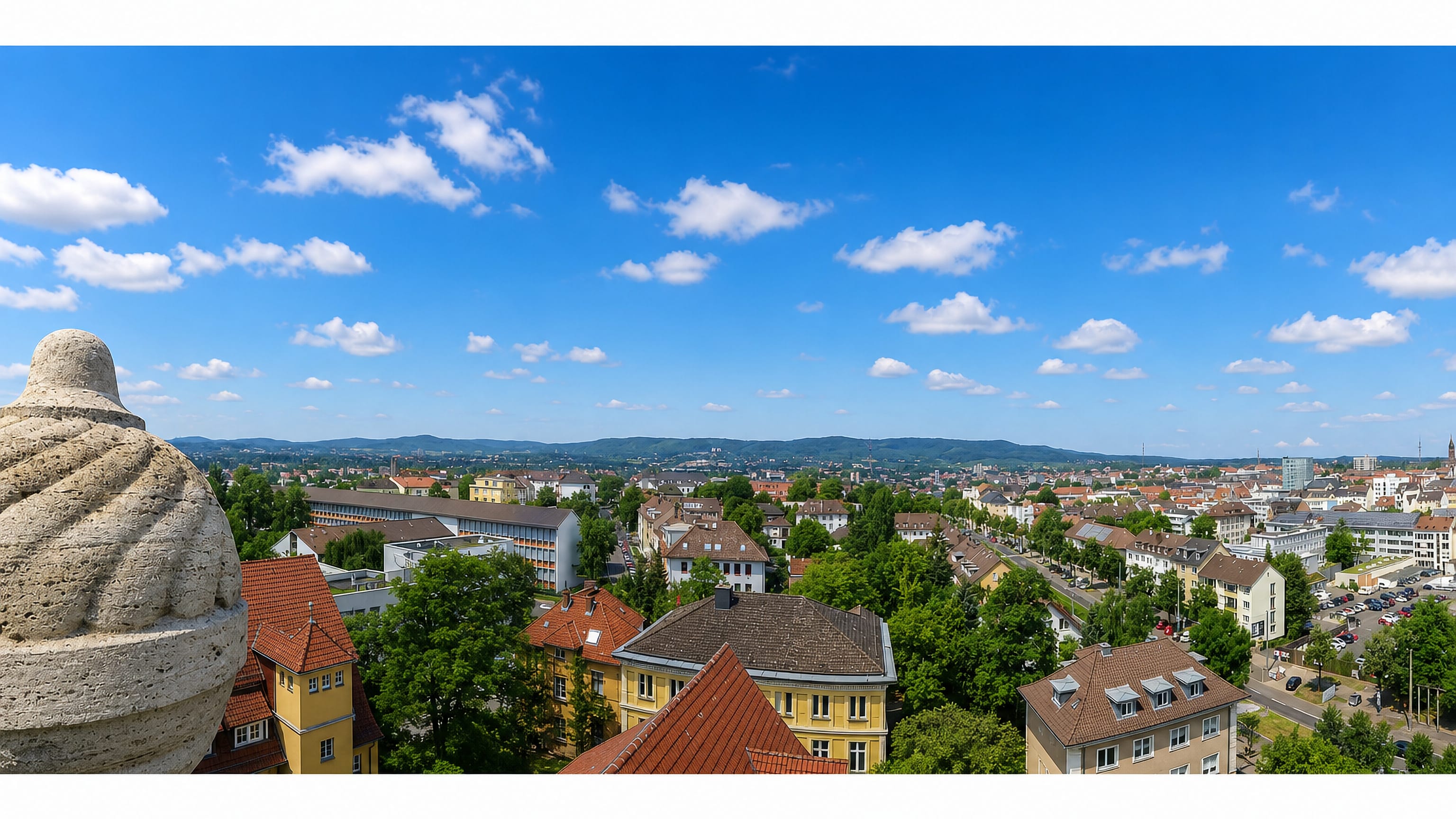 Panorama over Kassel's red rooftops and wooded hills viewed past a stone monument figure.