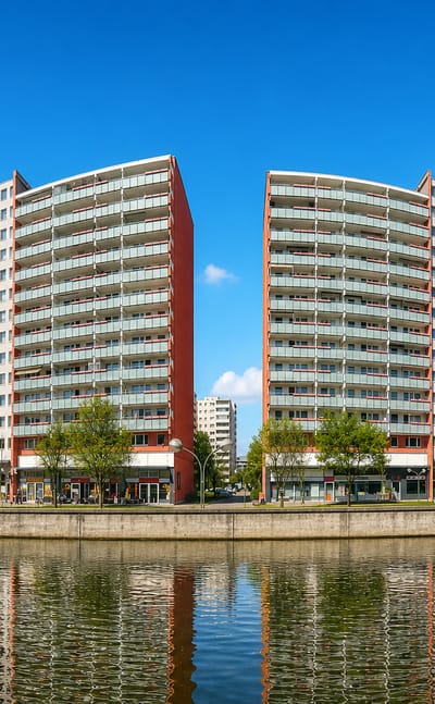 Twin curved red-and-white residential high-rises reflected in the Fennpfuhl lake, Berlin, with tower blocks behind
