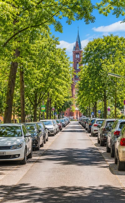 Tree-lined residential street of parked cars leading to a red-brick neo-Gothic church tower in Friedenau, Berlin.