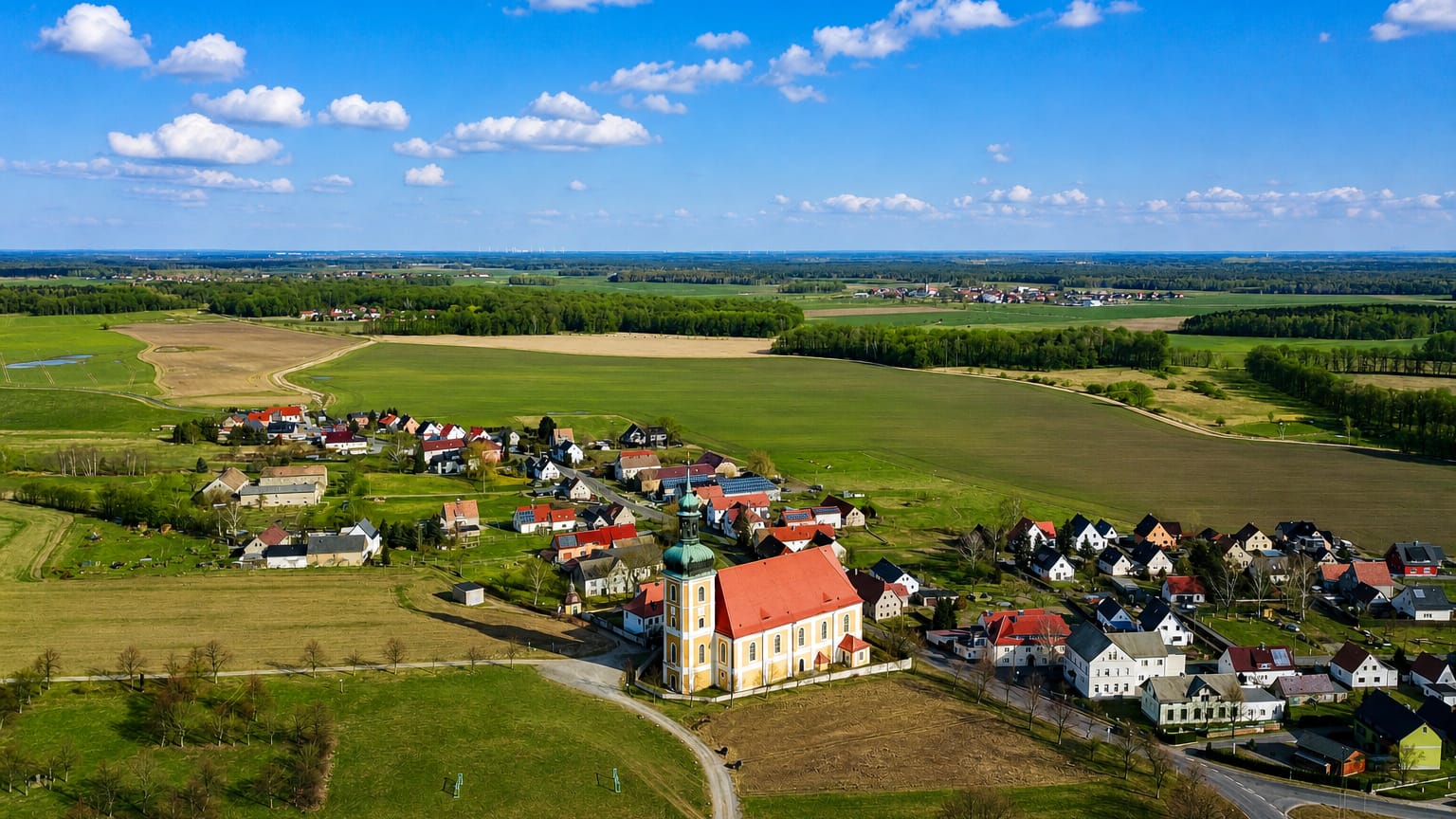 Aerial of a village with a baroque green-domed church amid open fields, evoking Rosenthal, Berlin