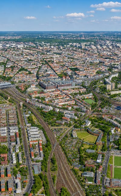 Aerial view over Rummelsburg in Berlin, showing the Rummelsburger See lake, converging railway lines and dense residential blocks stretching to the city horizon.