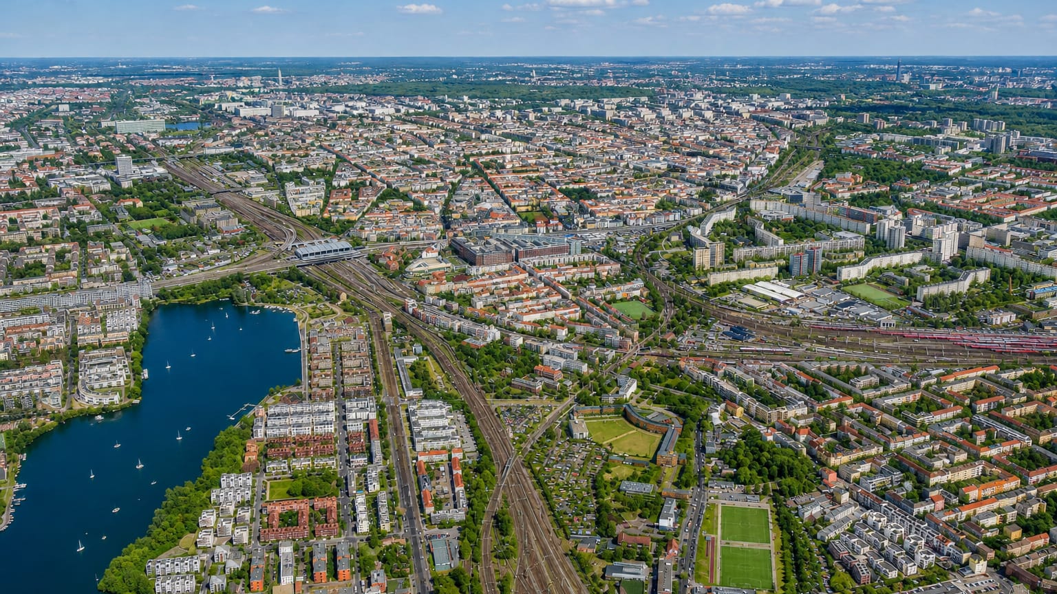 Aerial view over Rummelsburg in Berlin, showing the Rummelsburger See lake, converging railway lines and dense residential blocks stretching to the city horizon.