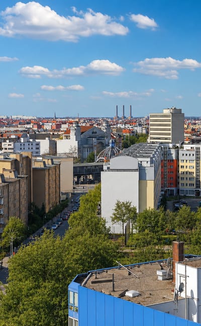 Rooftop view over mid-rise buildings, a red tower block and church spires in Schöneberg, Berlin, under a blue sky.