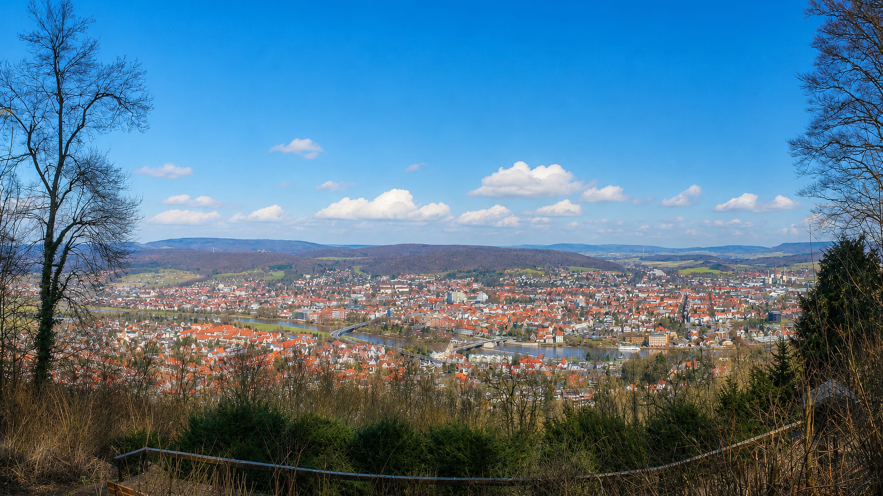 Panoramic hilltop view over Hameln's red-roofed old town and the River Weser, with the Weser Hills behind.