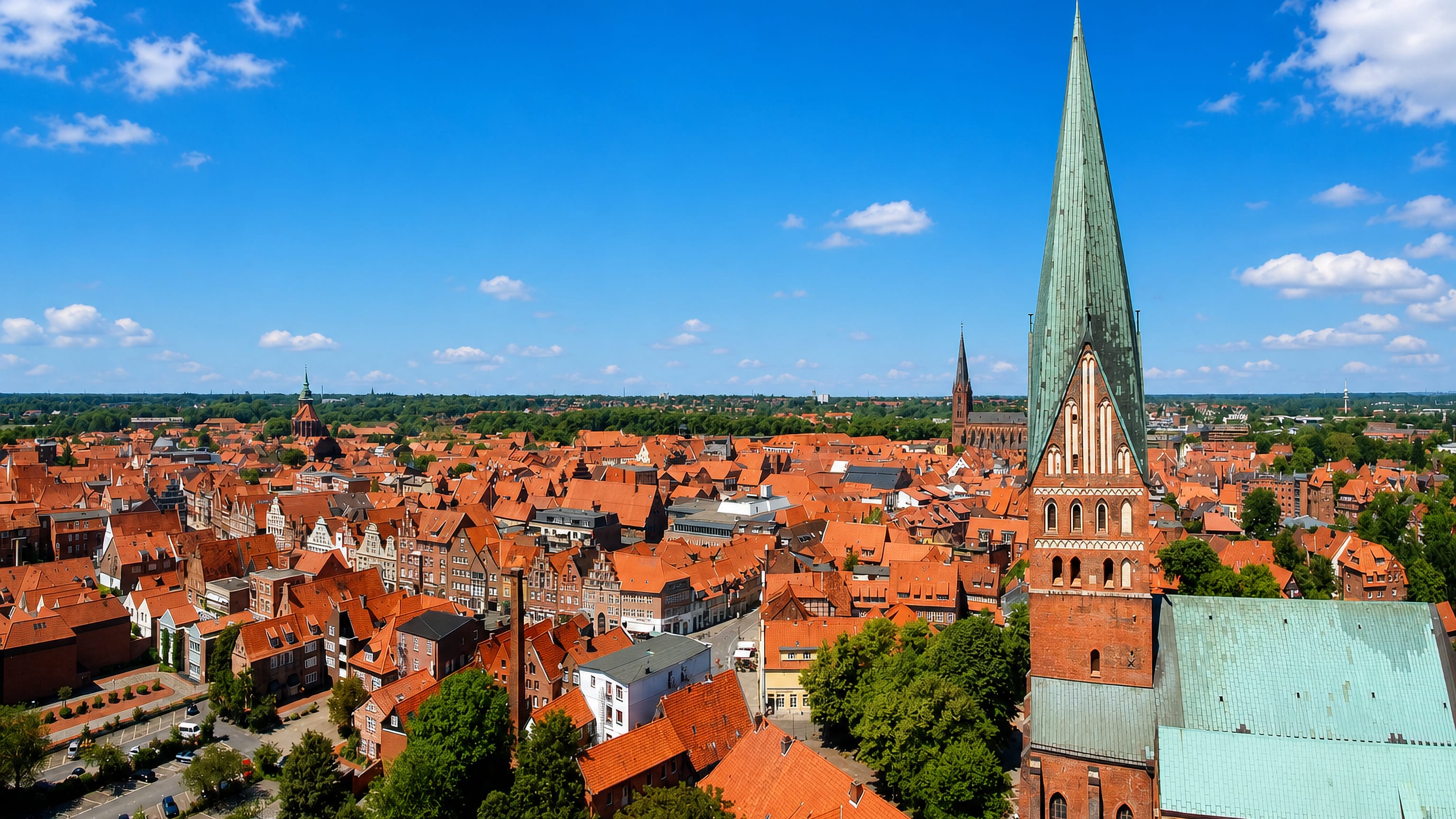 Lüneburg's red-tiled old town rooftops beneath the green copper spire of St John's Church under a blue sky.