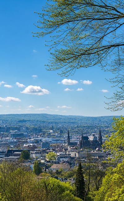 Aachen viewed from a wooded hillside, with the cathedral and town-hall towers rising above the city under a blue sky.