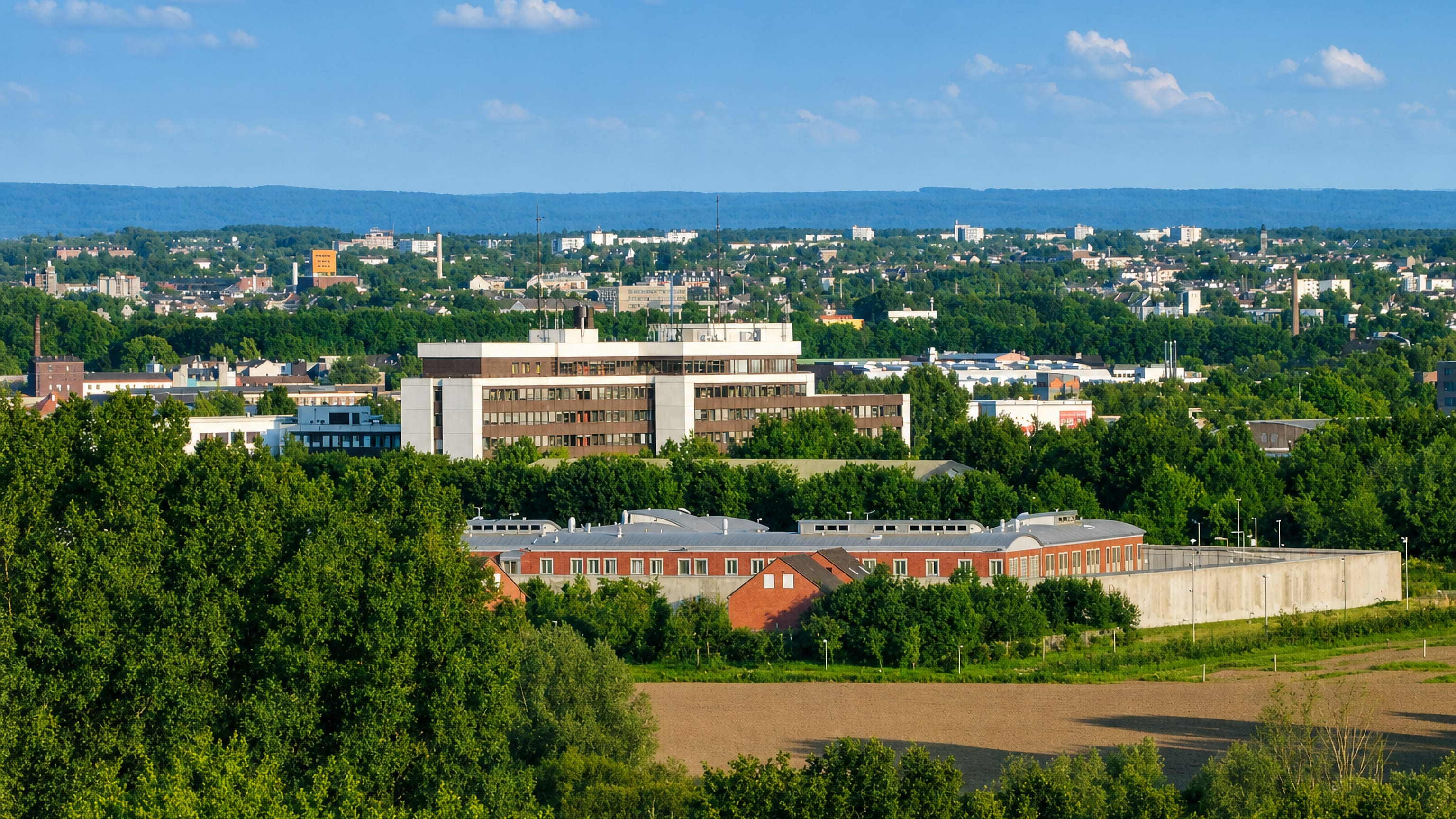 Green edge of Soers looking over Aachen's rooftops and wooded hills under a clear summer sky