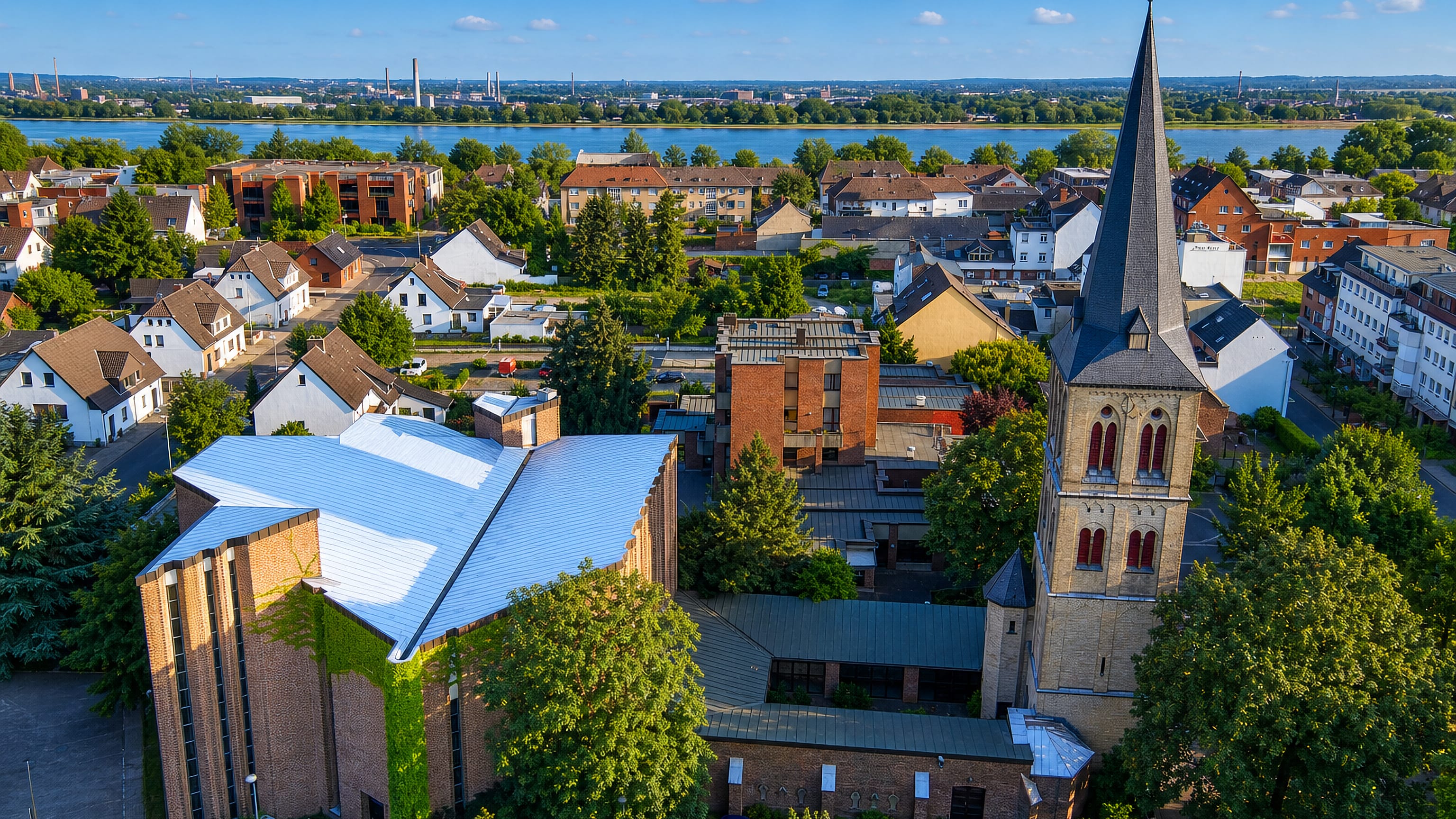 Church spire and rooftops of Dormagen, Germany, above the Rhine with industry on the far bank.
