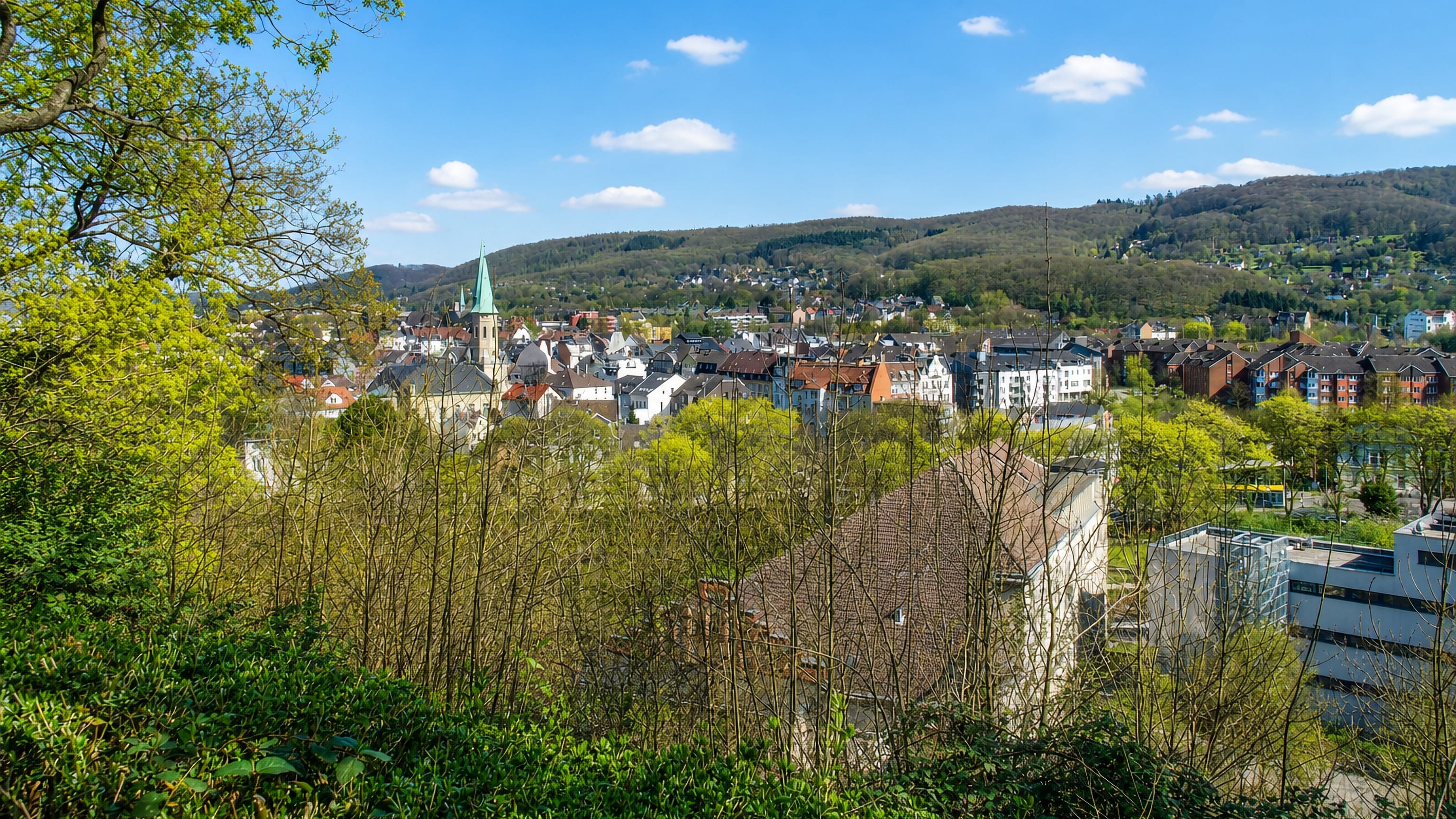 Haspe, Hagen: slate roofs and a green church spire in a wooded Sauerland valley seen from a hillside.