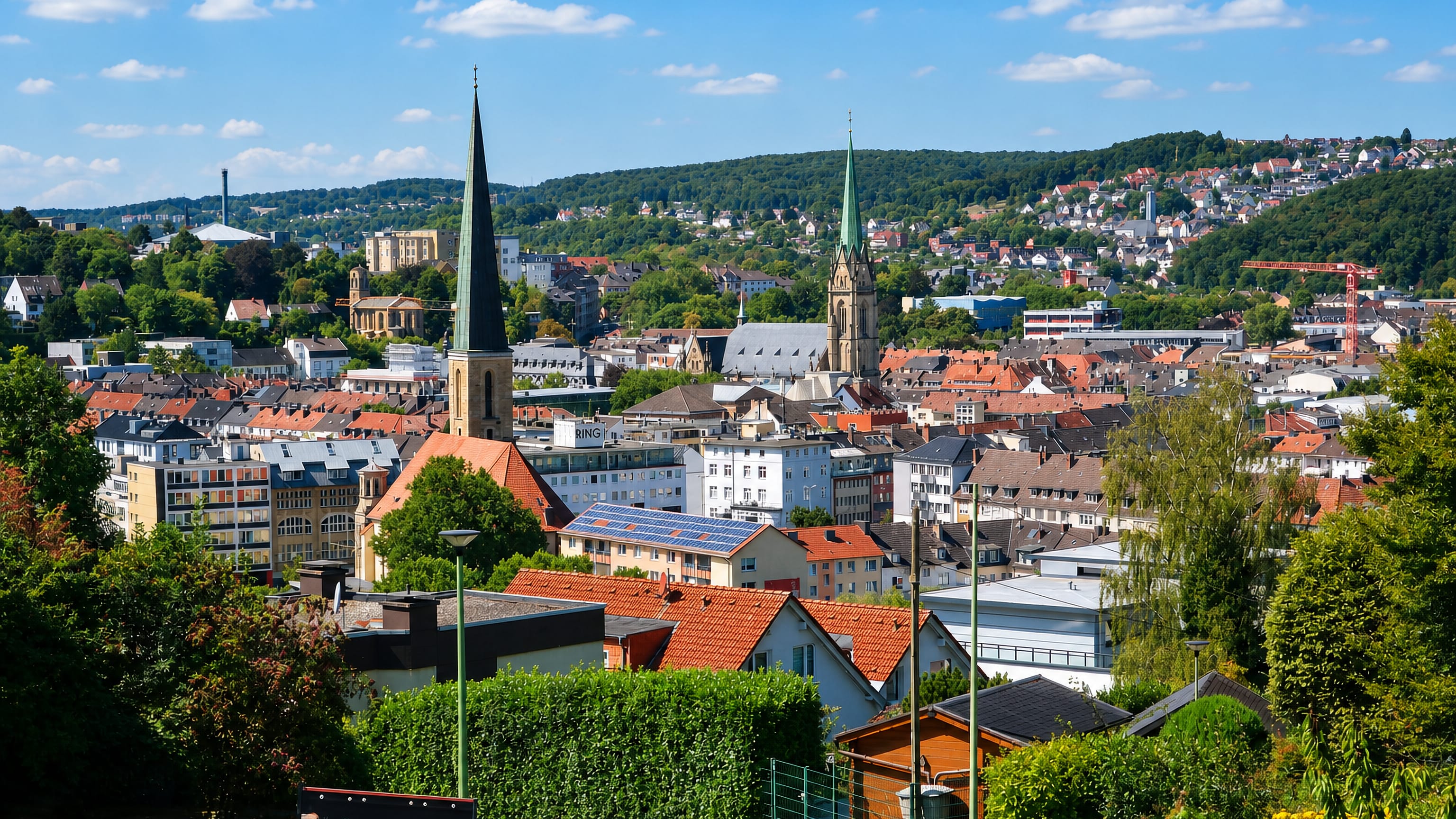 Daytime view across Hagen, Germany, with two church spires rising above red-tiled roofs and wooded hills behind.