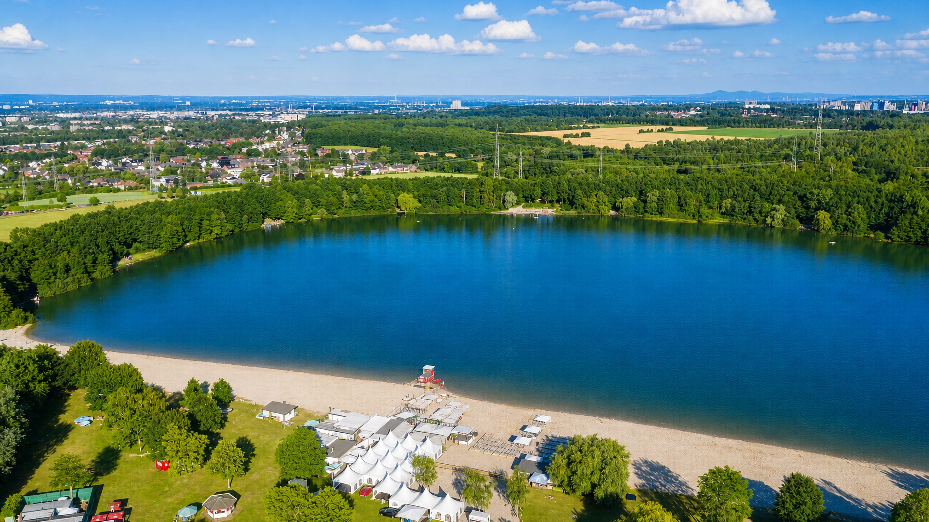 Aerial view of a lake beach and wooded shore at Huerth near Cologne, with the Rhineland skyline beyond.