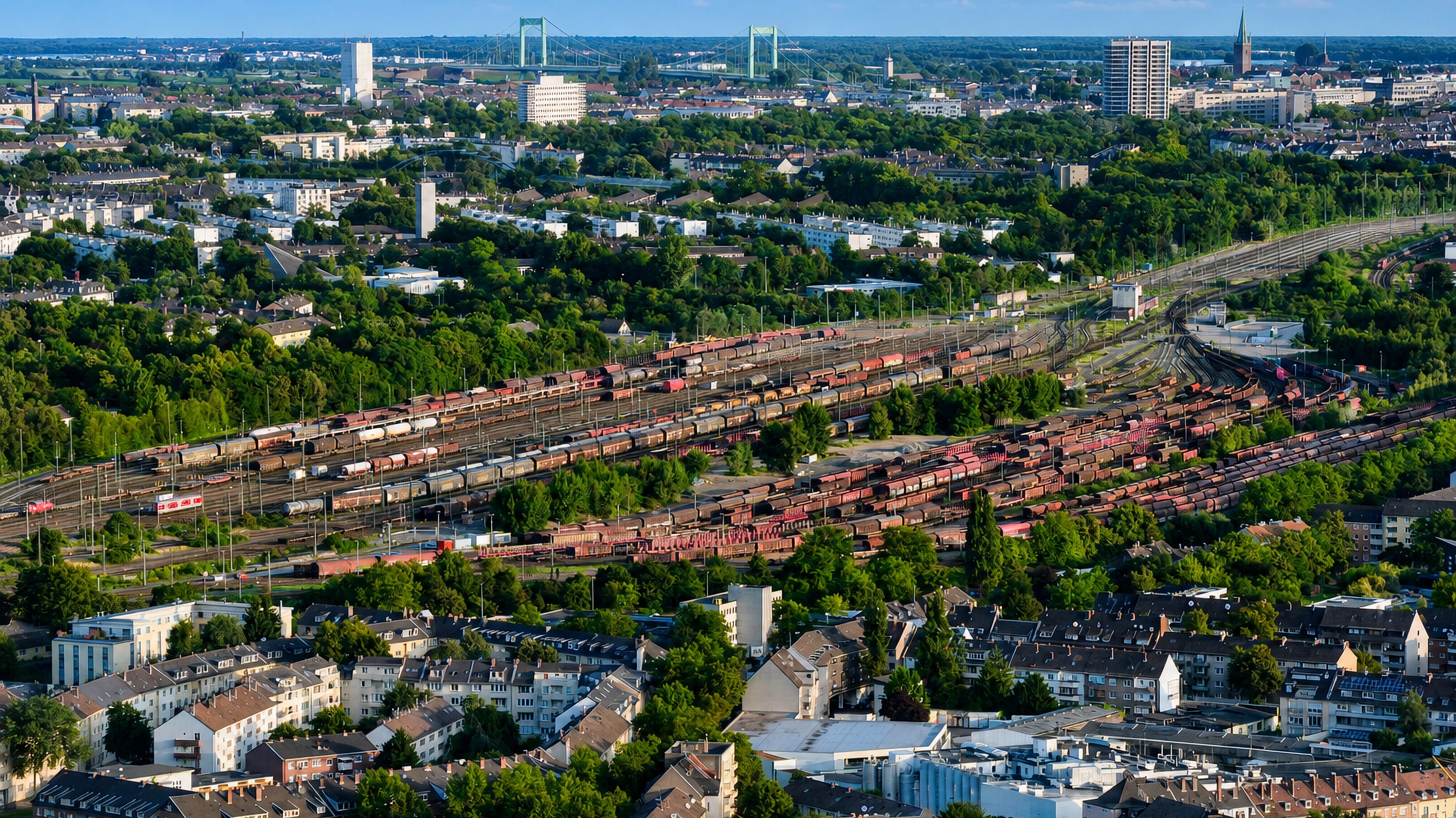 Aerial over Buchforst's rail freight yard and rooftops with a Rhine bridge on the Cologne horizon.