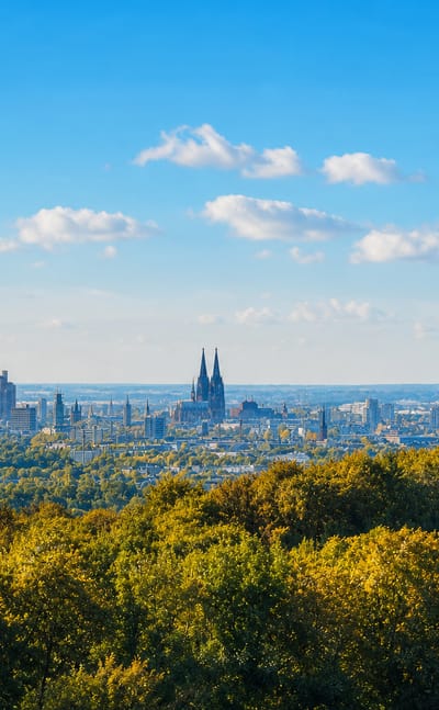 Cologne skyline across autumn treetops, with Cologne Cathedral's twin spires and the Colonius tower under a blue sky.