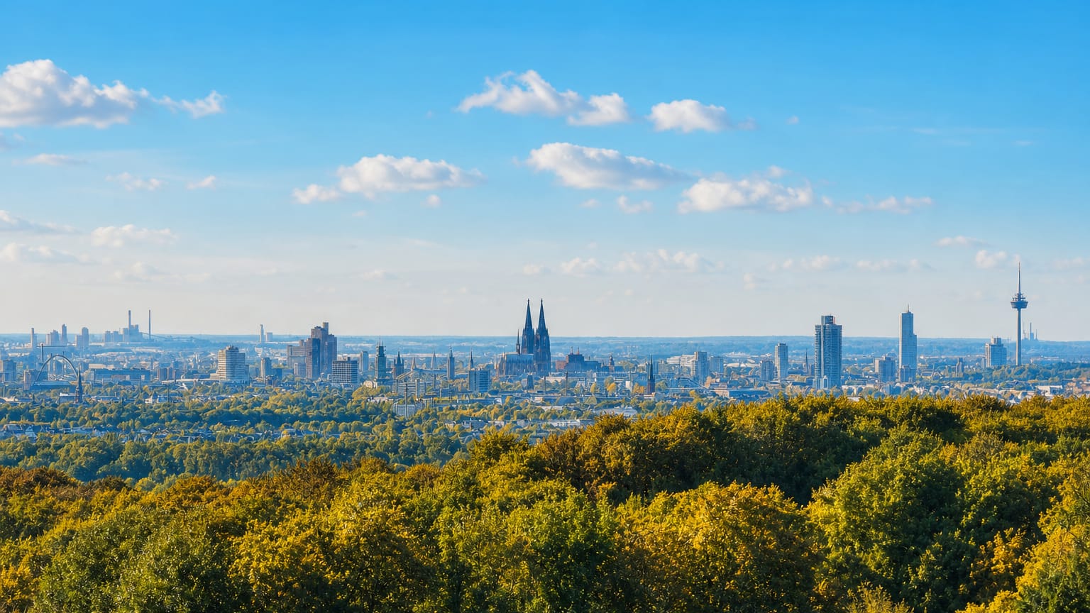 Cologne skyline across autumn treetops, with Cologne Cathedral's twin spires and the Colonius tower under a blue sky.