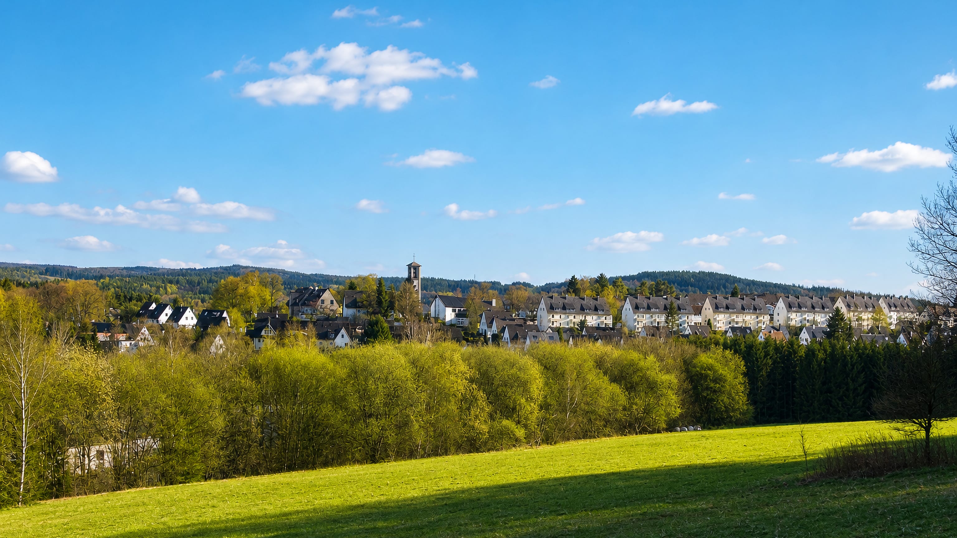 Slate-roofed houses and a church tower on a wooded Sauerland ridge above meadows near Lüdenscheid, Germany.