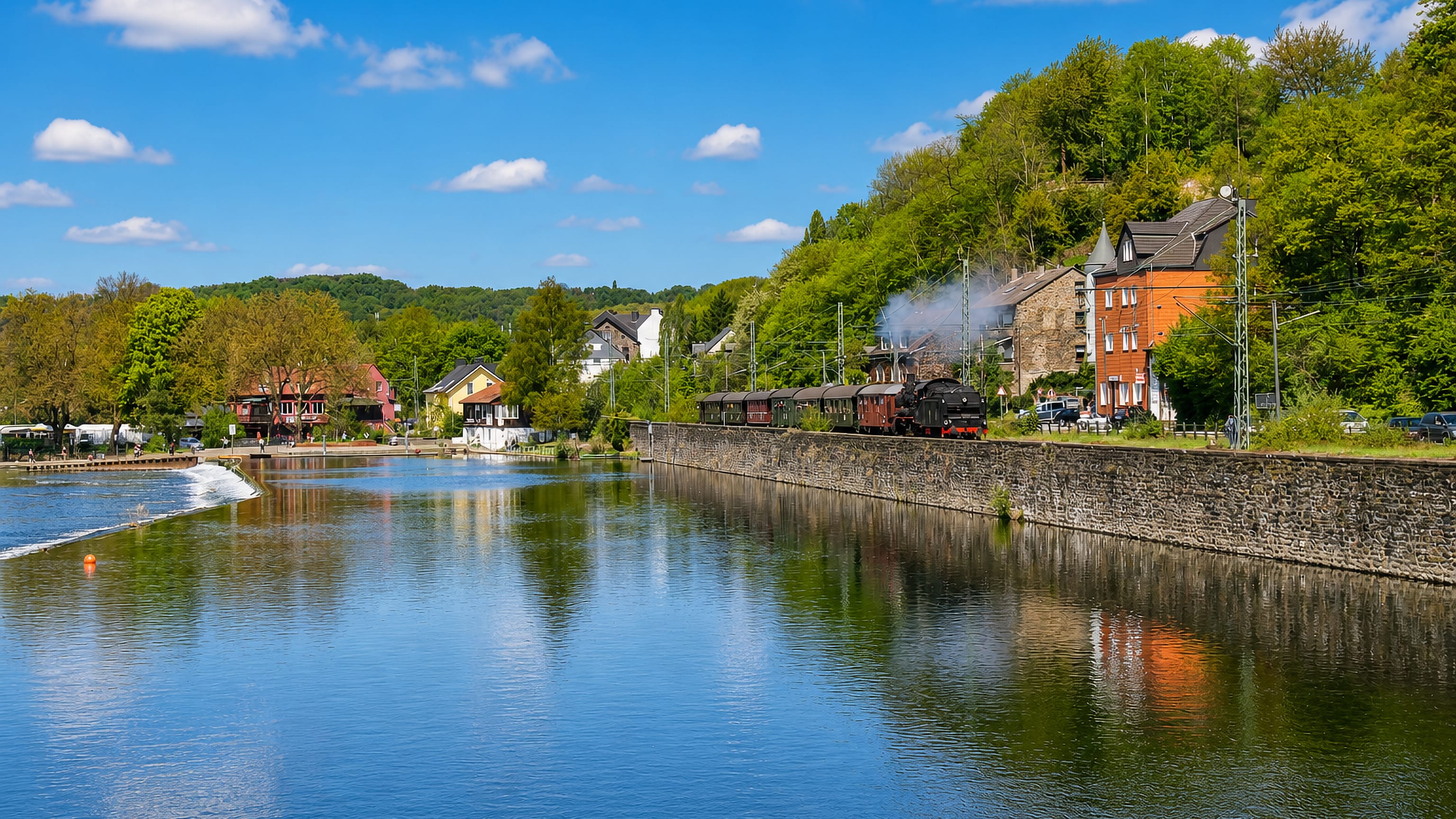 A steam train running along the Ruhr embankment below wooded hillside houses in Dahlhausen.
