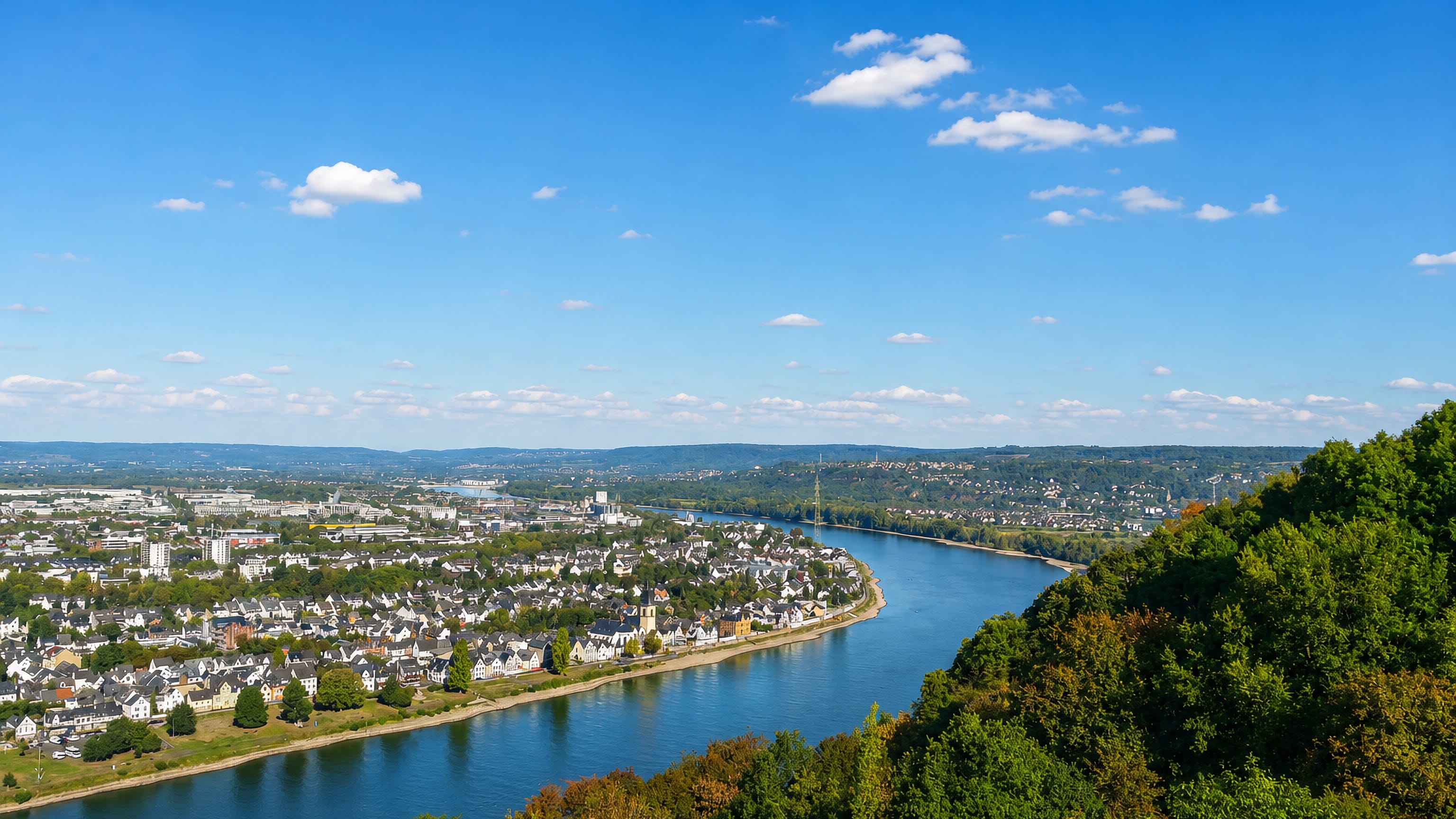 A town spread along a bend of the Rhine river with wooded hills beyond, near Koblenz, Germany.