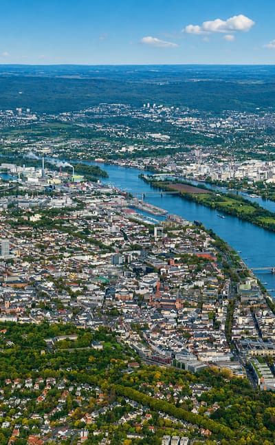 Aerial view over Mainz with old-town rooftops and boats along the wide River Rhine under a blue sky.