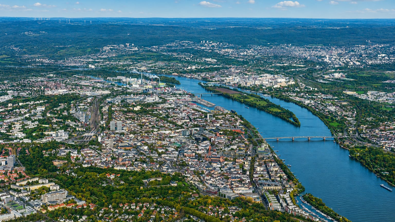 Aerial view over Mainz with old-town rooftops and boats along the wide River Rhine under a blue sky.