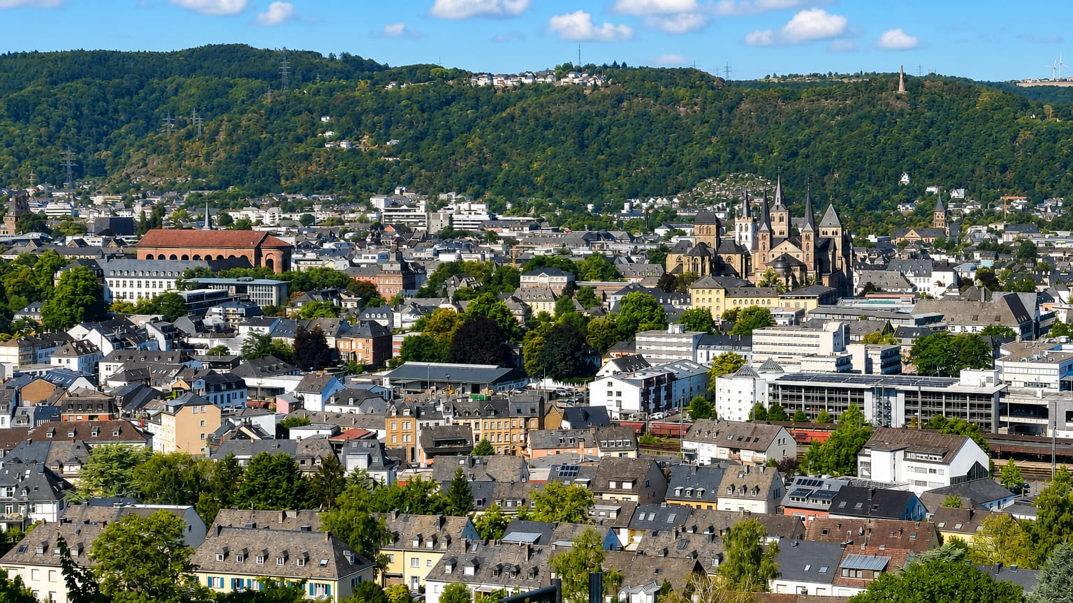 Trier's rooftops and the twin-towered cathedral below wooded hills under a blue sky.