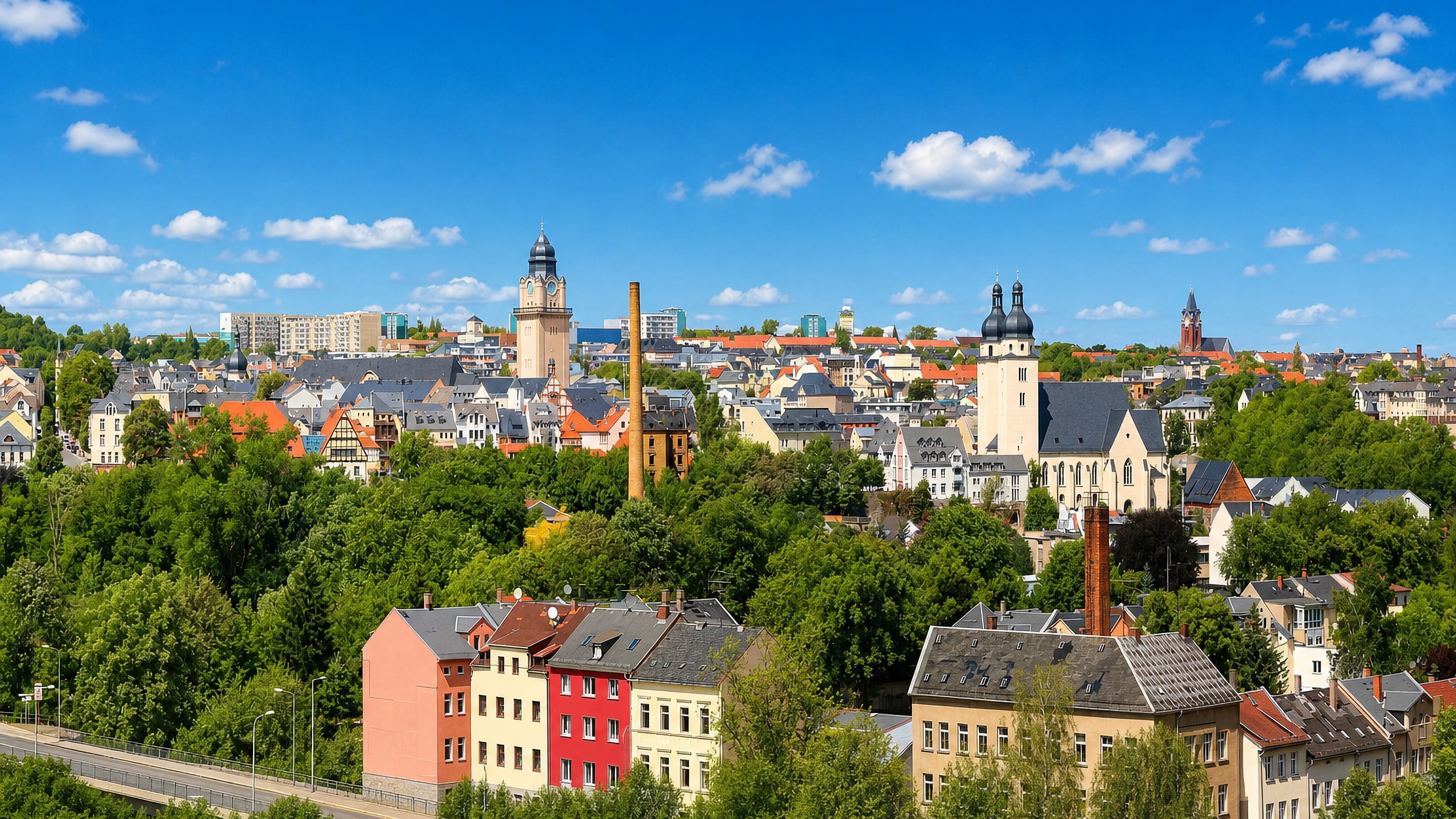 Plauen rooftops below the old town hall tower, the twin-domed Johanniskirche and a brick mill chimney amid wooded hills.