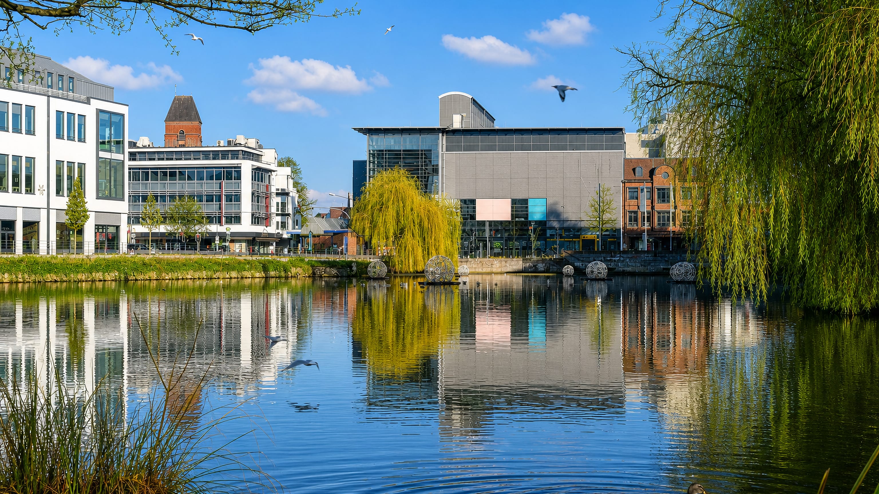 Neumunster, Germany: modern town-centre buildings and willows mirrored in a still town pond on a clear spring day