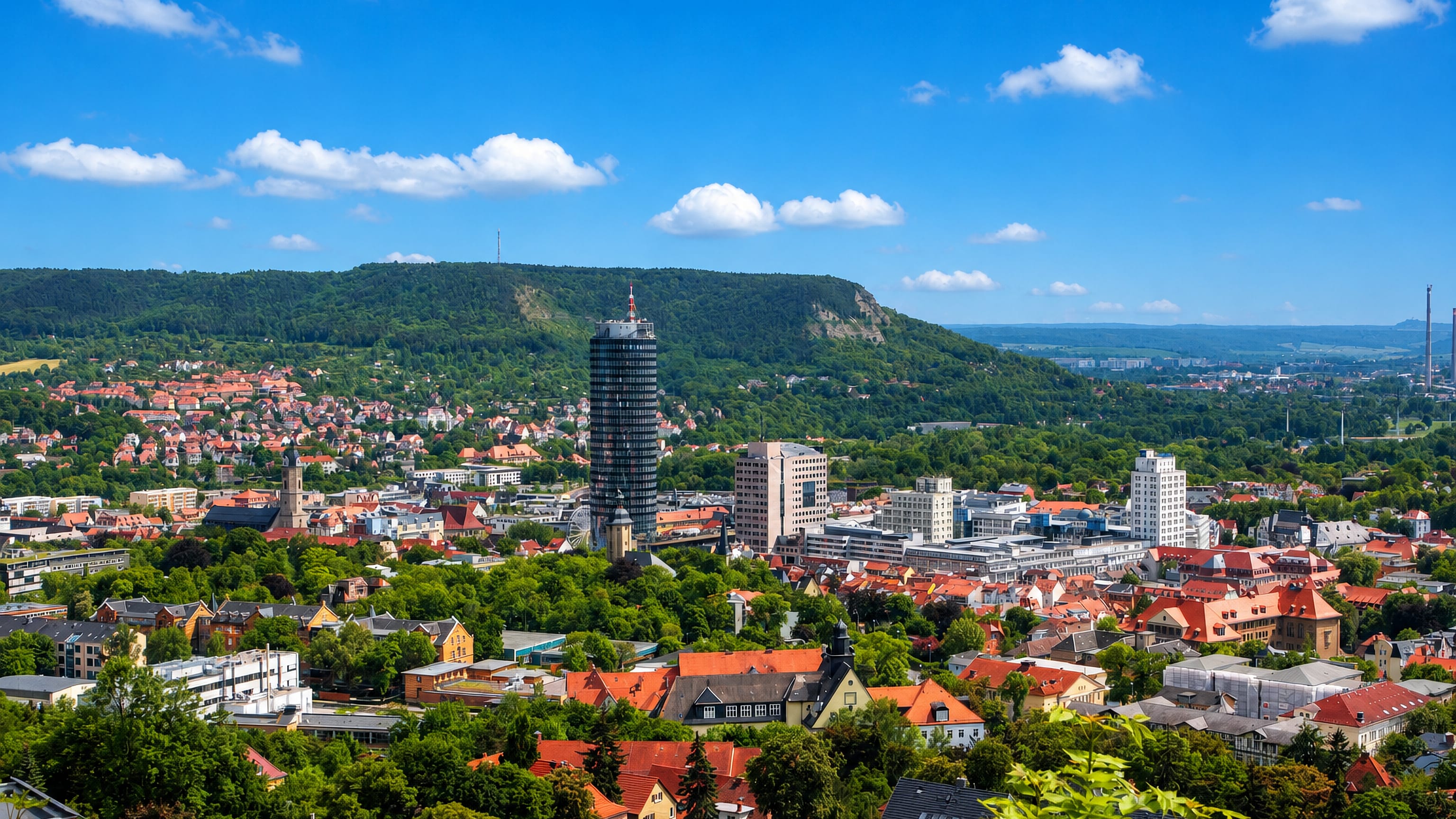 Jena's cylindrical JenTower rising above red-roofed houses in the Saale valley below wooded hills.