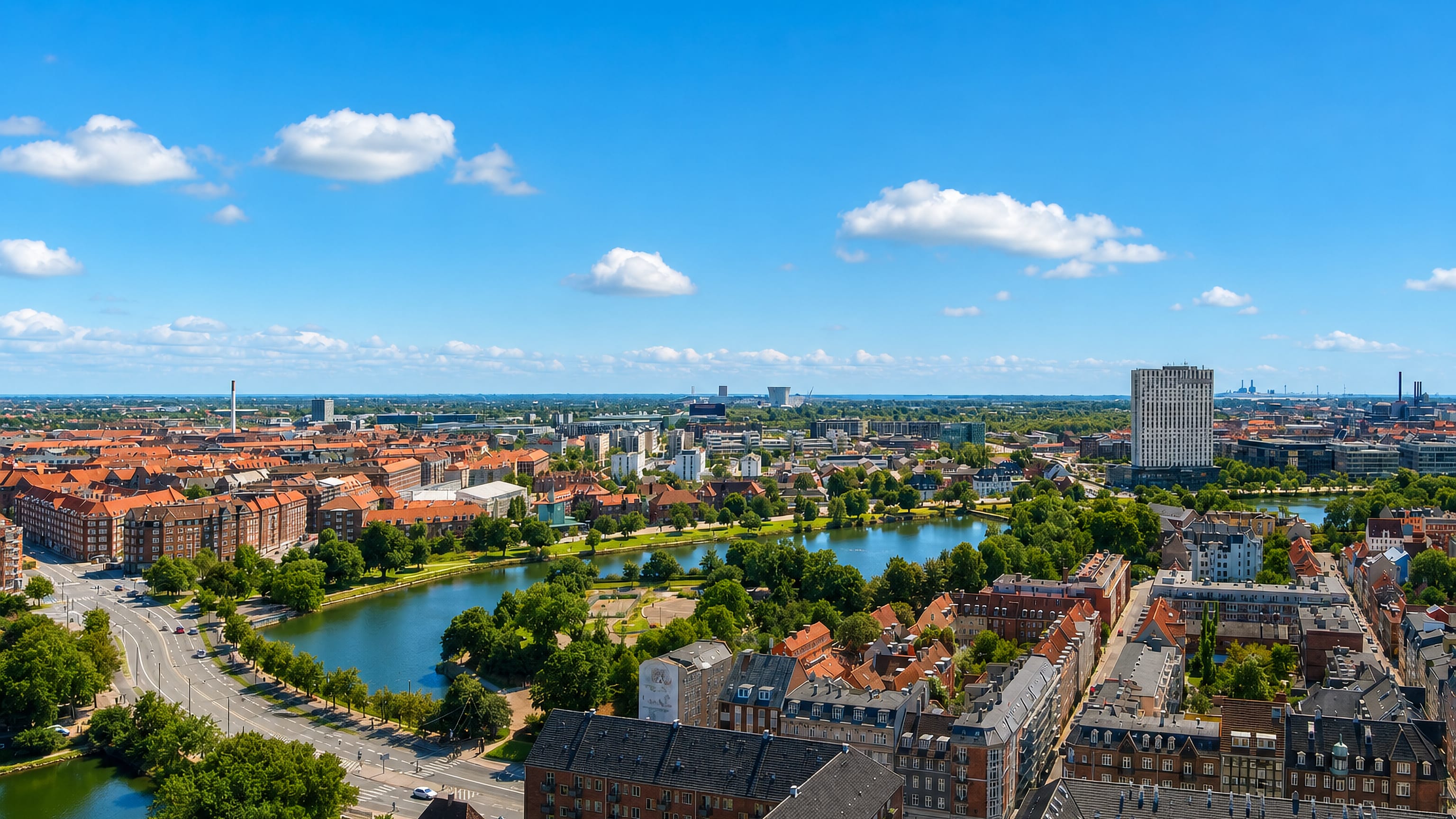 Aerial over Amagerbro's red-roofed blocks and a tree-lined lake toward Copenhagen's harbour chimneys