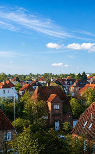 Red-tiled rooftops of traditional houses among green trees in Valby, Copenhagen, beneath a clear blue sky with a distant tower