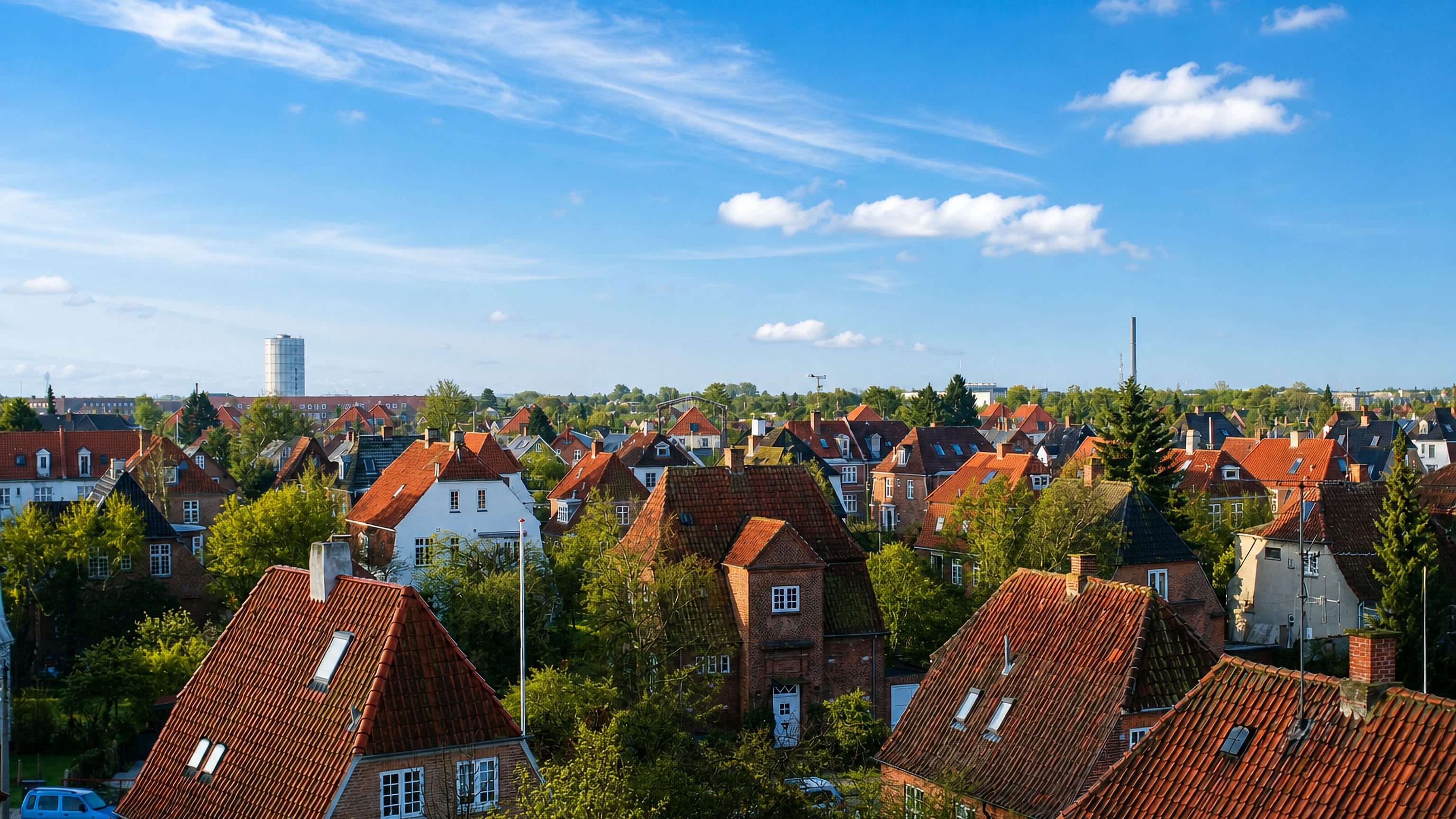 Red-tiled rooftops of traditional houses among green trees in Valby, Copenhagen, beneath a clear blue sky with a distant tower