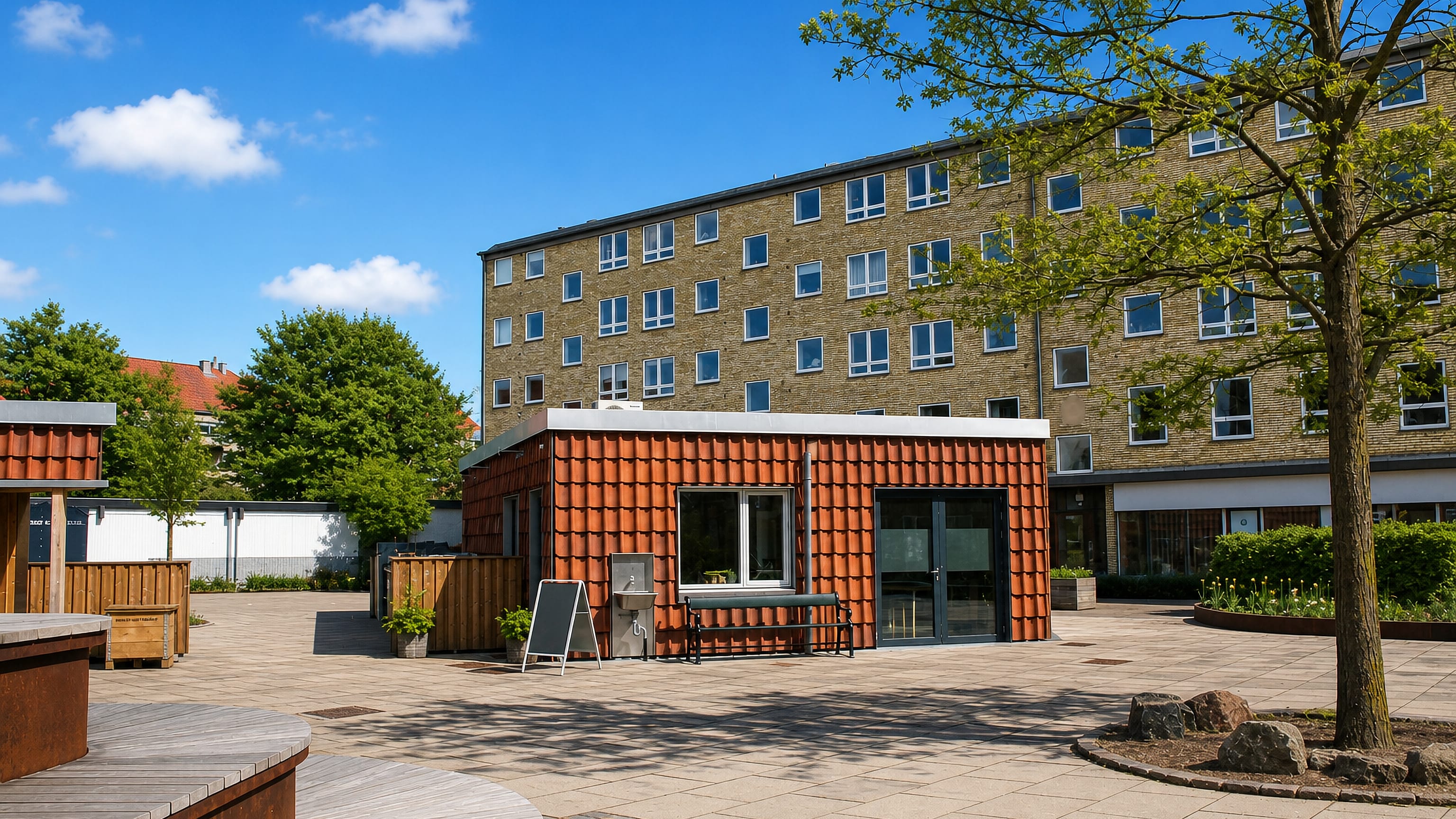 Sunlit paved square in Vanløse, Copenhagen, with a tile-clad pavilion and yellow-brick apartment blocks