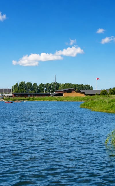 Frederikssund marina on Roskilde Fjord, with moored sailboats, a Danish flag and reed-lined shore under a bright blue sky