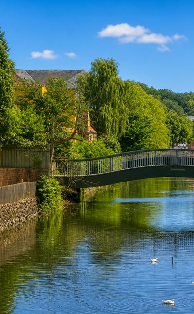 A green arched footbridge over the canal in Frederiksvaerk, Denmark, with a yellow building and swans on the water.